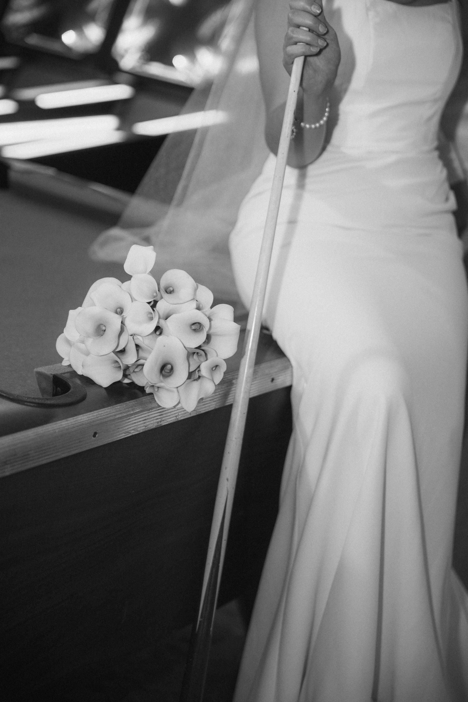 Bride in white gown holding a pool cue beside a bouquet of calla lilies on a billiard table, black and white photo.