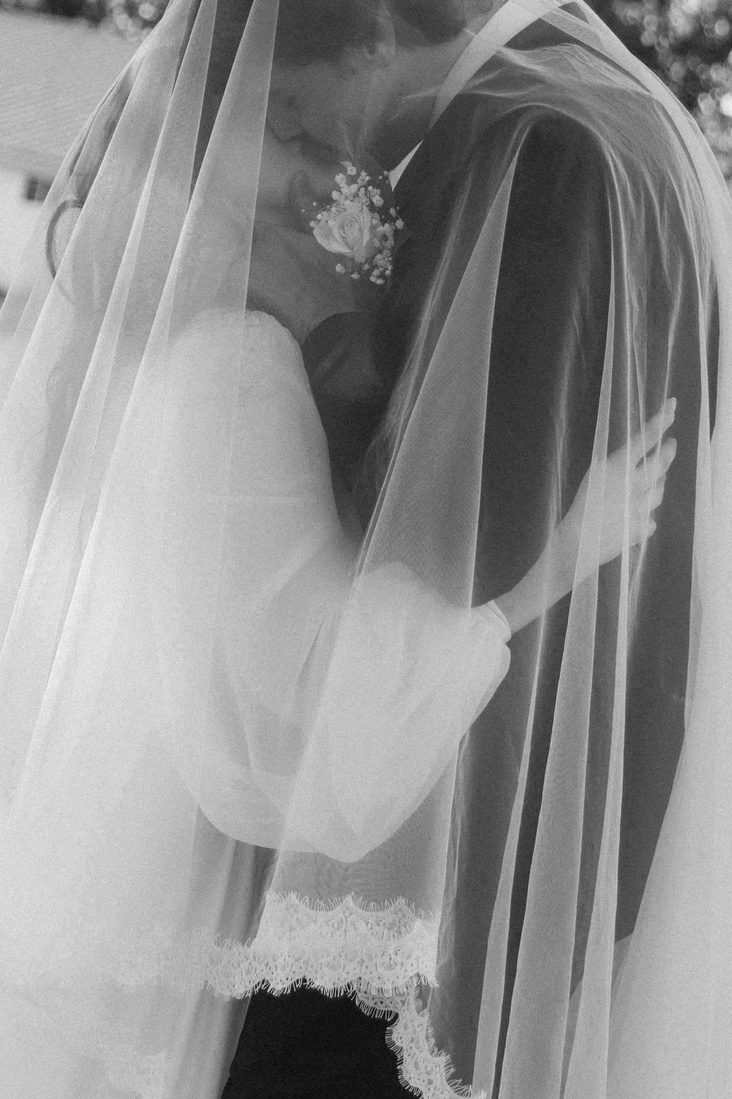 Black and white photo of bride and groom embracing under a flowing veil, with floral boutonniere visible.