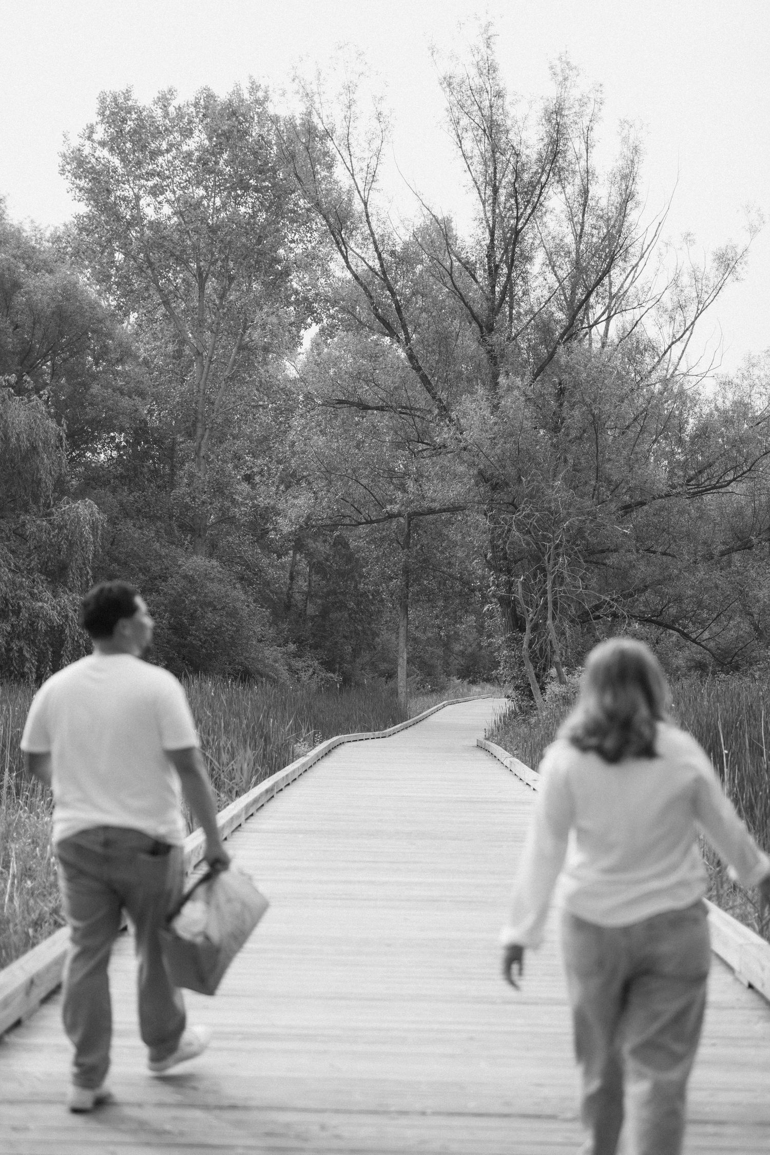 Black and white photo of two people walking on a wooden boardwalk path through a lush forest park.