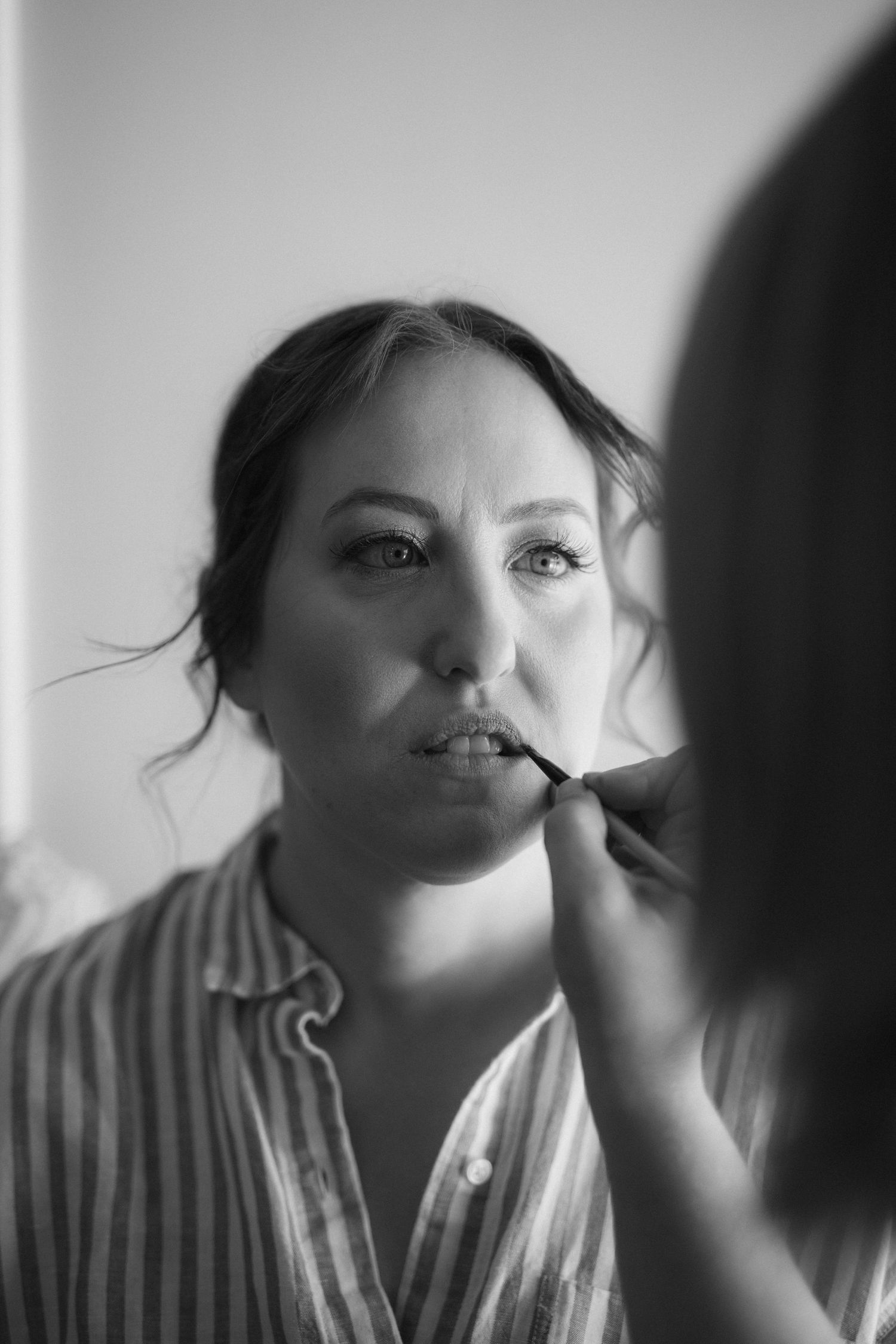 Black and white photo of makeup being applied to someone wearing a striped button-up shirt.