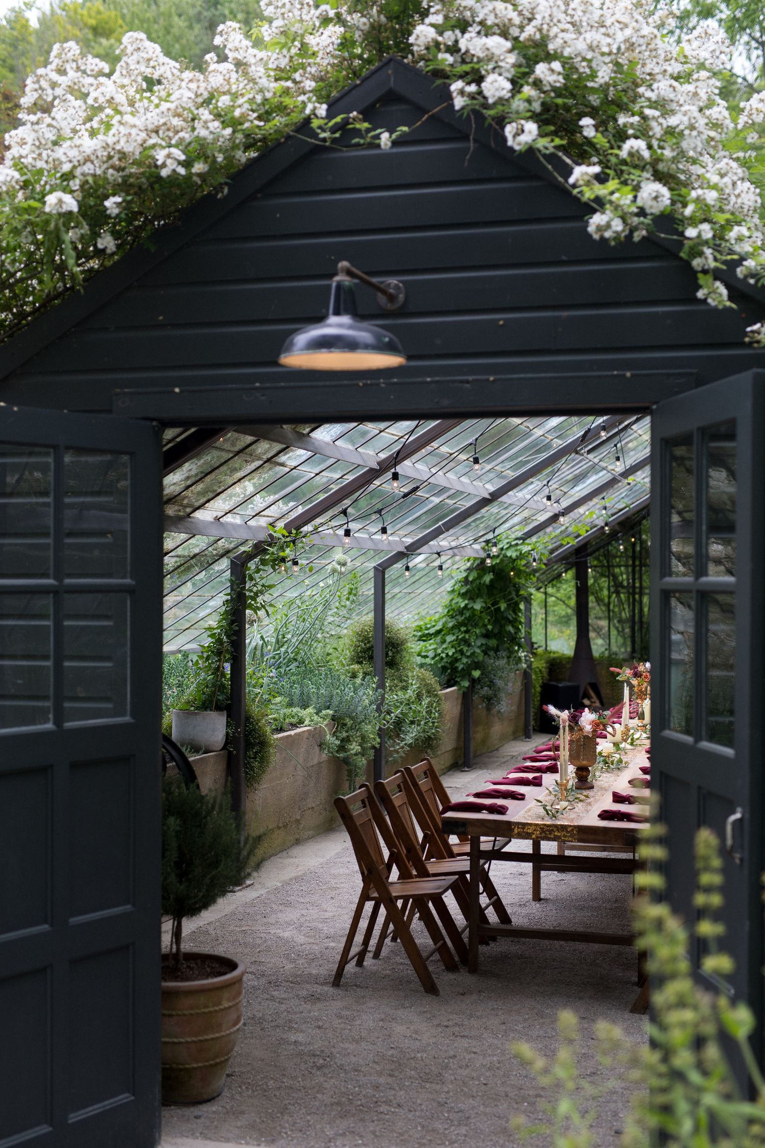 A rustic greenhouse dining area with wooden tables and chairs under flowering vines and glass roof panels.
