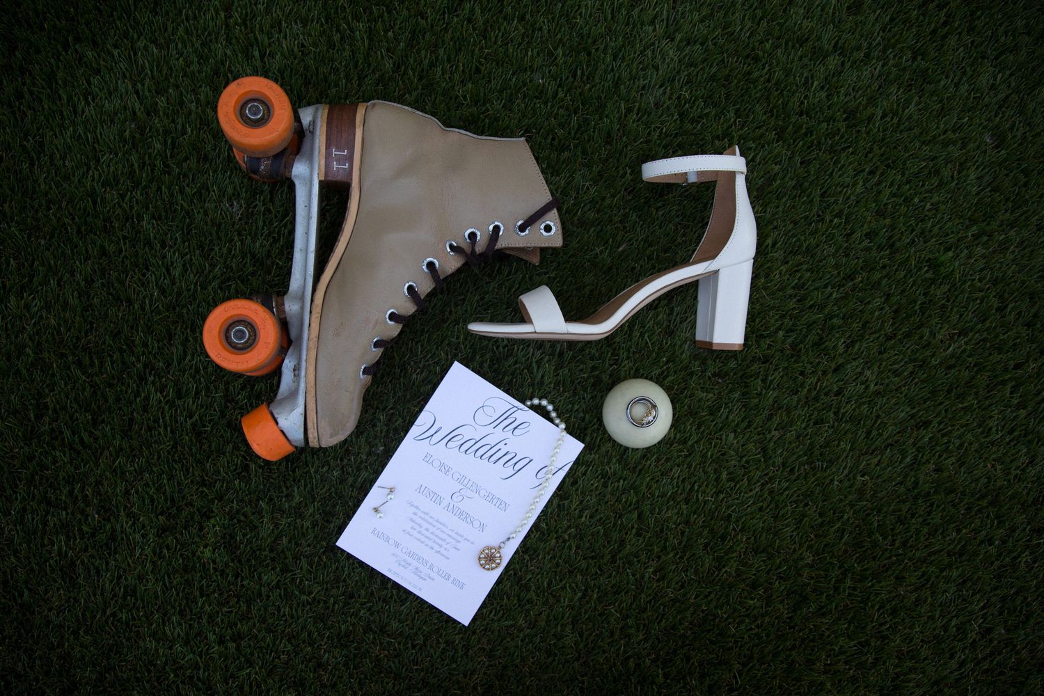 Vintage roller skates with orange wheels beside a white heeled sandal and wedding invitation on green grass.