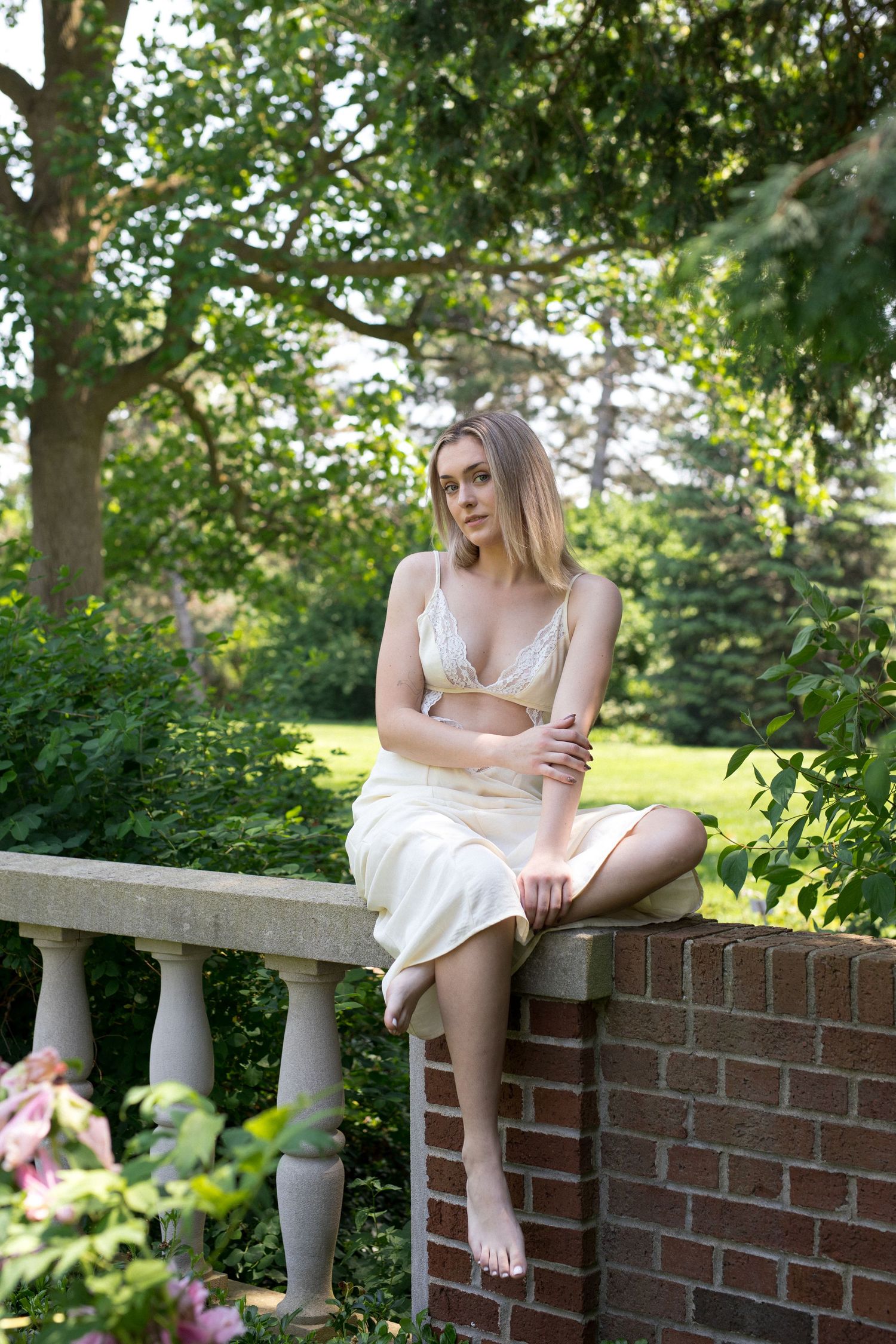 A person in a white dress sits on a brick wall balustrade in a sunny garden setting.