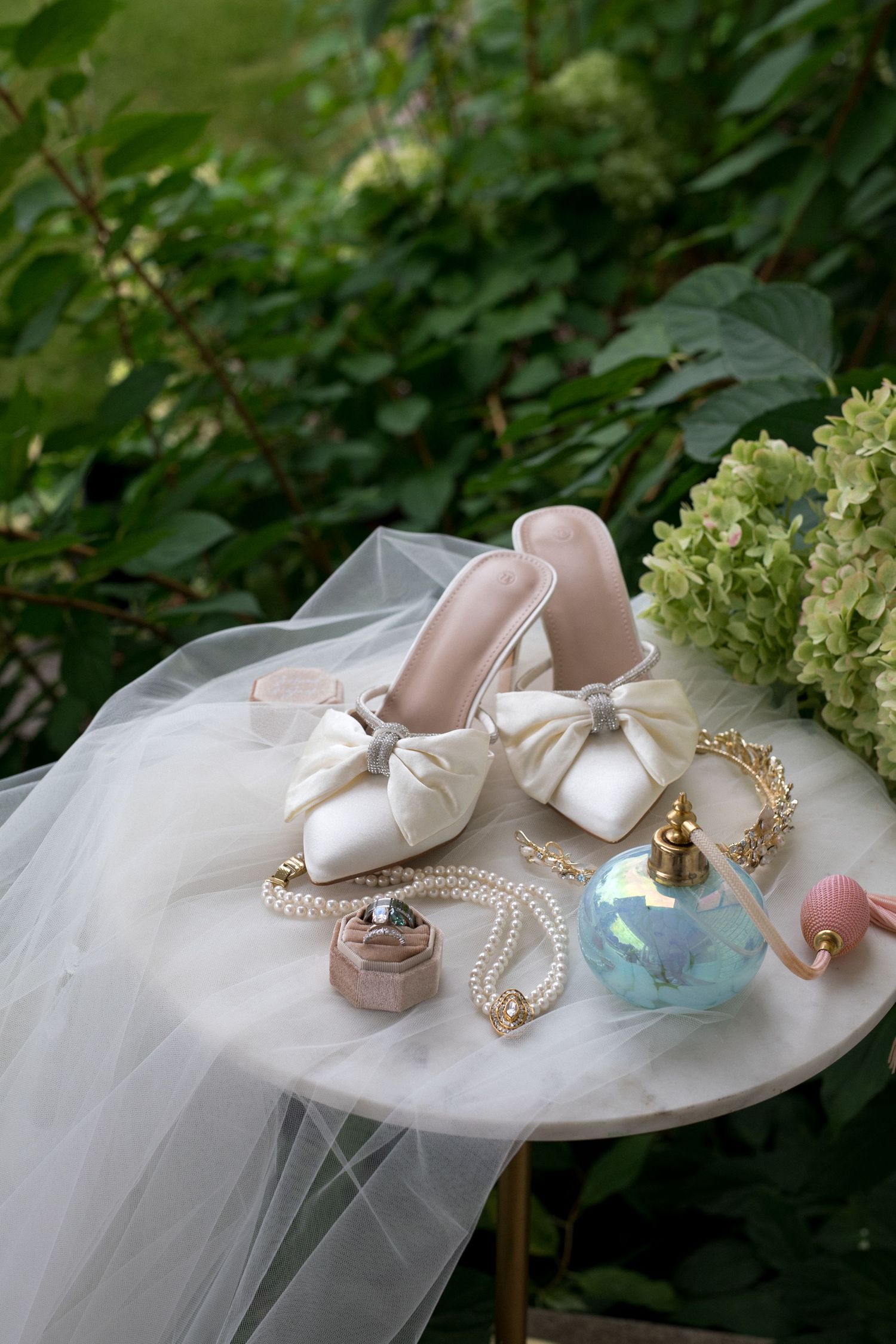 Wedding accessories including rings, pearl jewelry, and satin shoes elegantly arranged on white tulle with greenery backdrop.