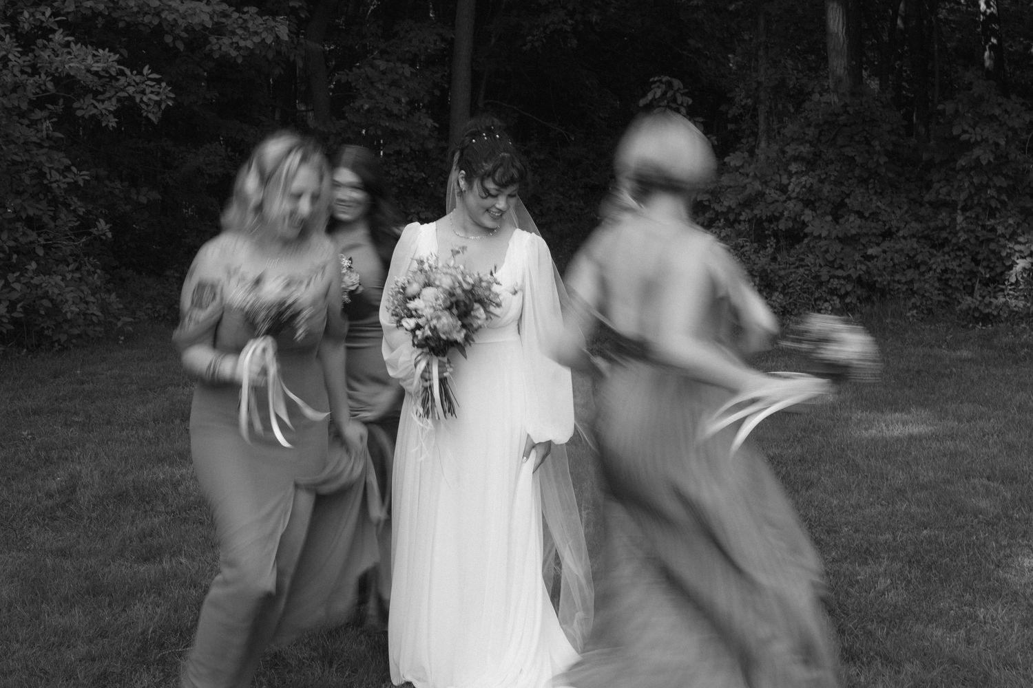 A black and white artistic motion blur photograph of a wedding party dancing outdoors in a garden setting.