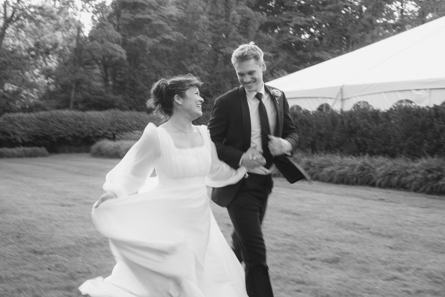 A bride and groom walking together on a grassy field with trees in the background.