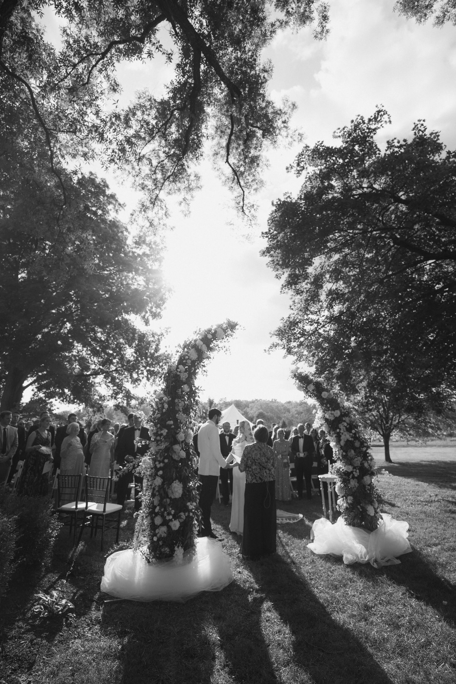 A black and white wedding ceremony silhouette with guests gathered under trees during golden hour lighting.