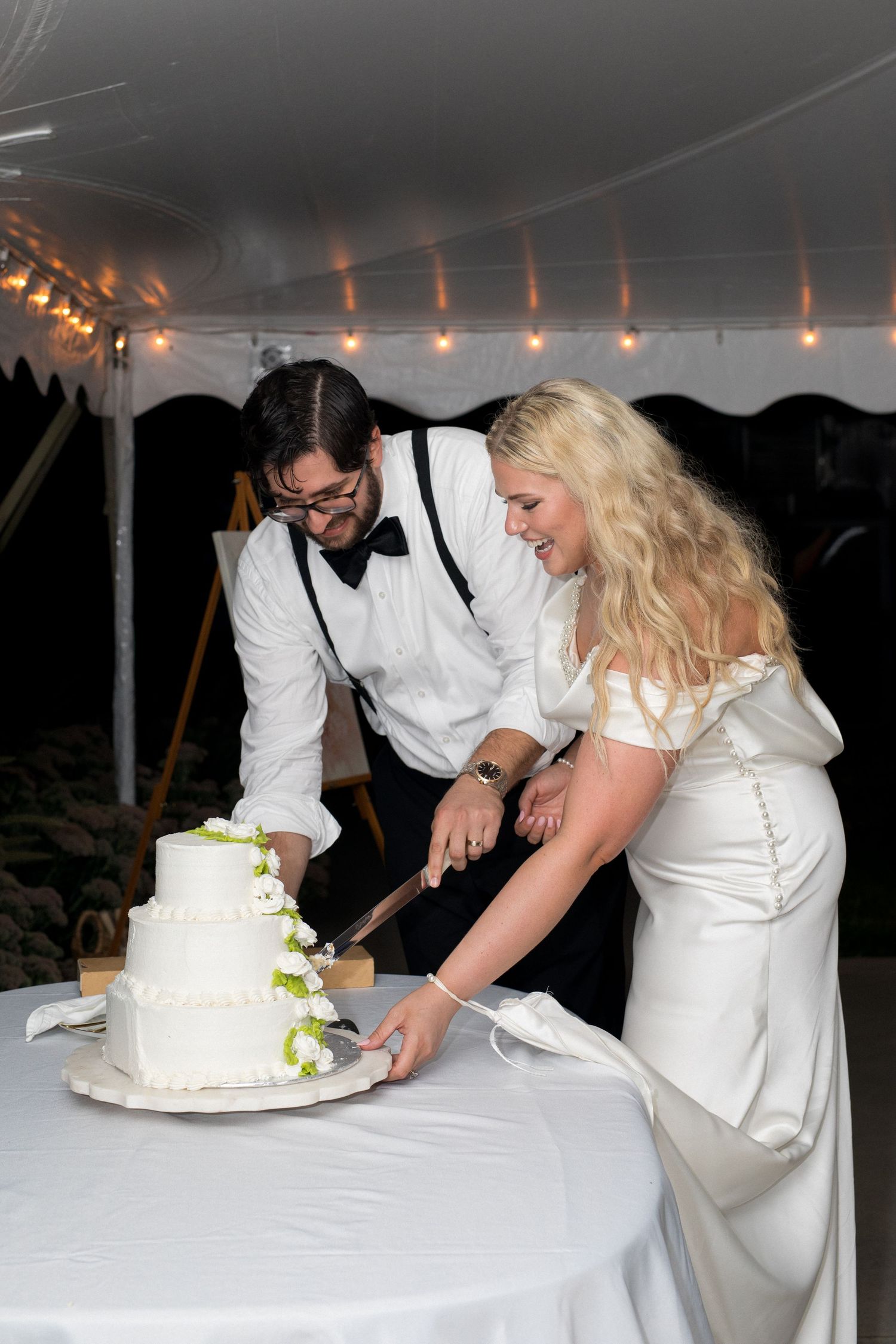 A couple in white wedding attire cuts their tiered white wedding cake together at an evening reception.