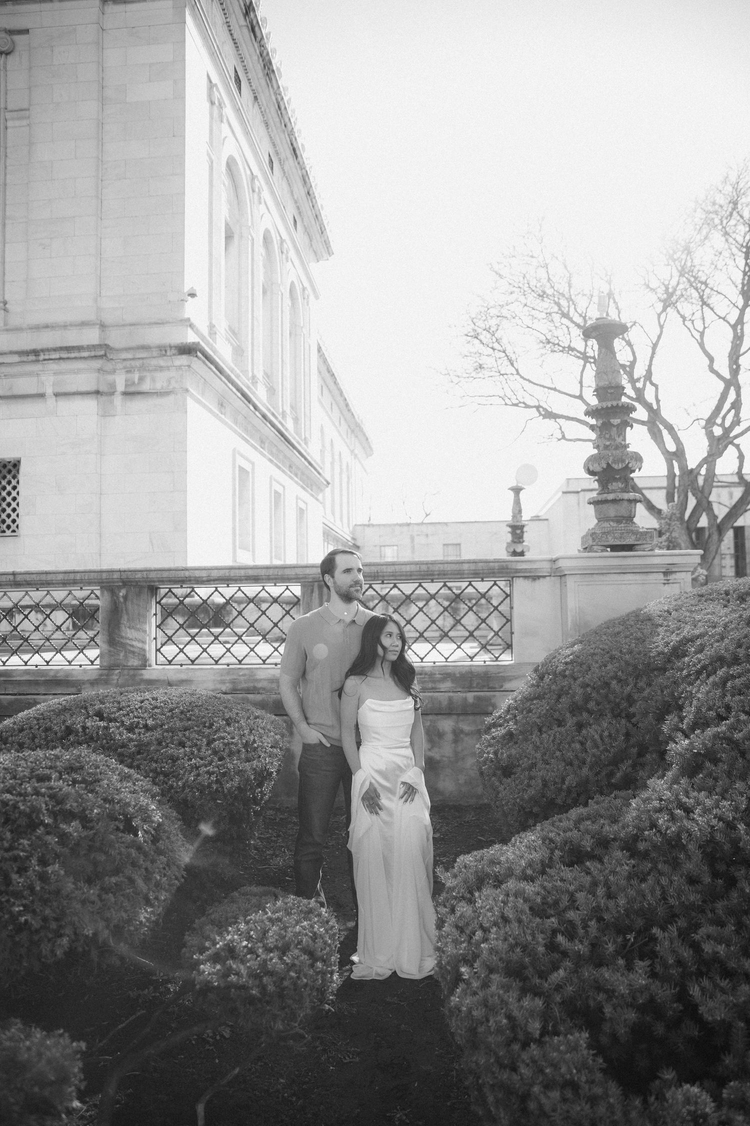 Couple posing in formal garden with manicured hedges beside elegant building, black and white photo.
