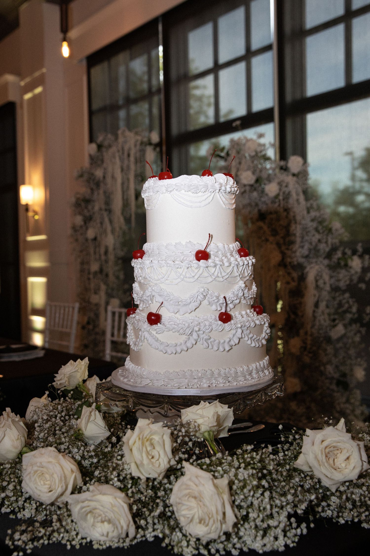 Elegant four-tier white wedding cake with red cherries and ruffled frosting, surrounded by white roses and baby's breath.