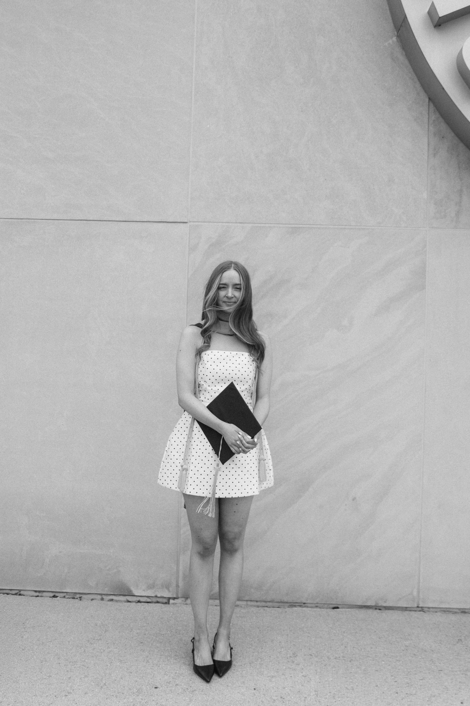 Young woman in polka dot strapless mini dress holding clutch purse, posing outdoors in black and white photo.