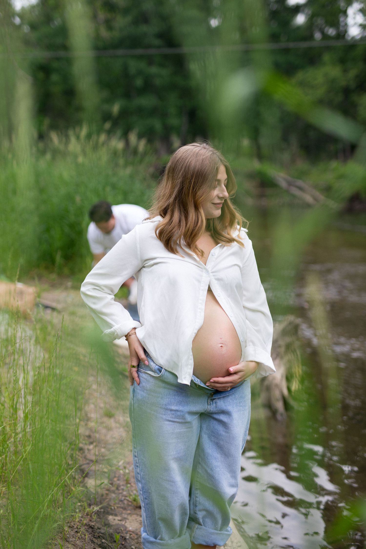 Pregnant woman in white shirt and jeans cradles her baby bump outdoors near a serene creek surrounded by lush greenery.