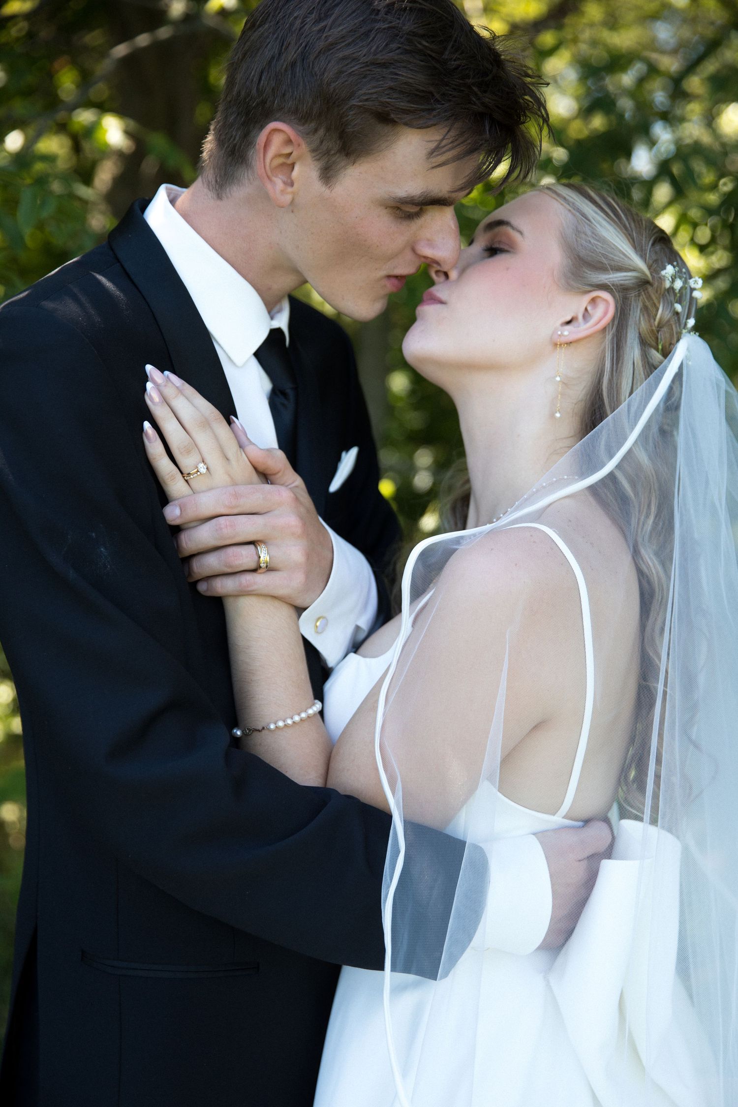 A romantic wedding couple shares a tender kiss outdoors with sunlit trees in the background.