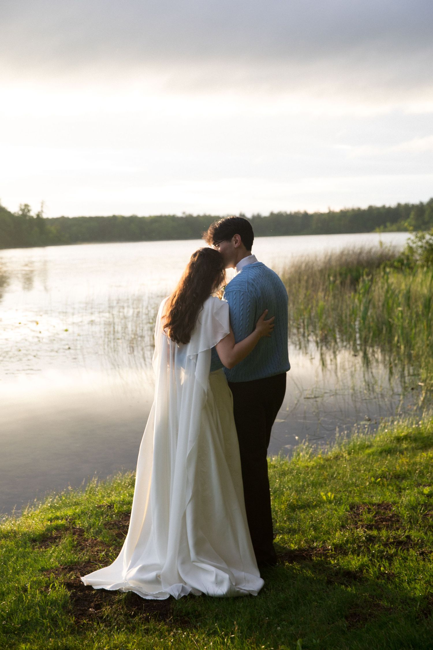 A couple in wedding attire shares a romantic moment by a tranquil lake at sunset, surrounded by lush greenery.