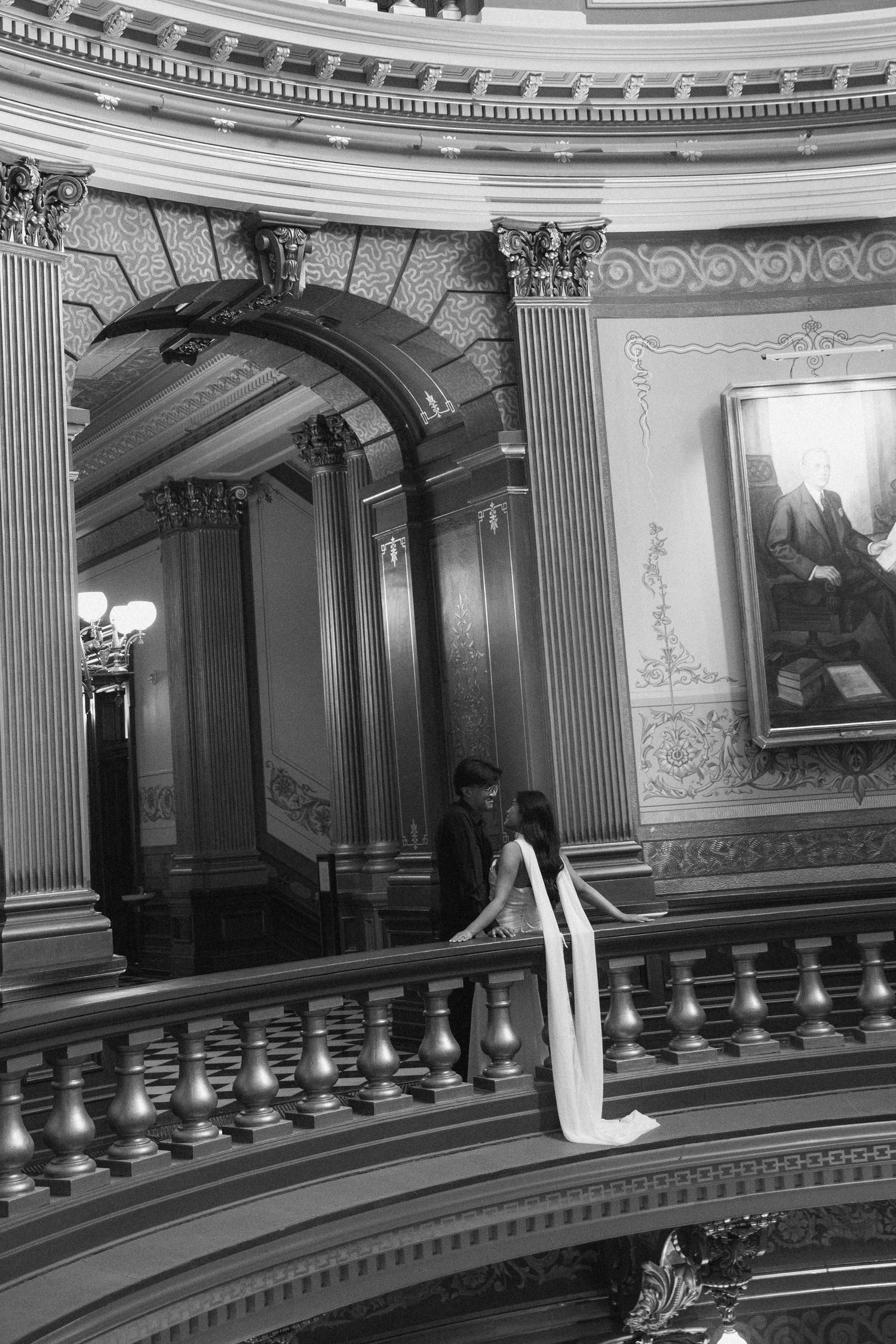 Black and white photo of a couple on an ornate balcony inside a grand historic building with classical architecture.