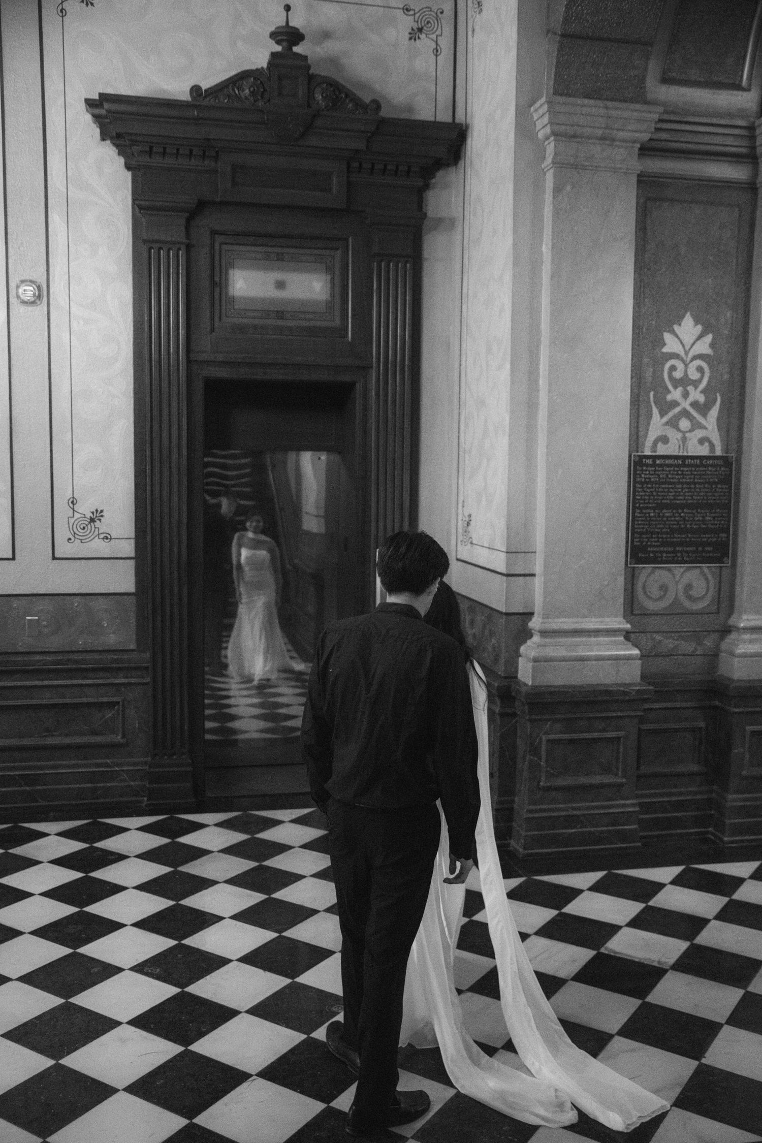 Black and white photo of groom standing before ornate doorway with bride's reflection visible on checkered floor.