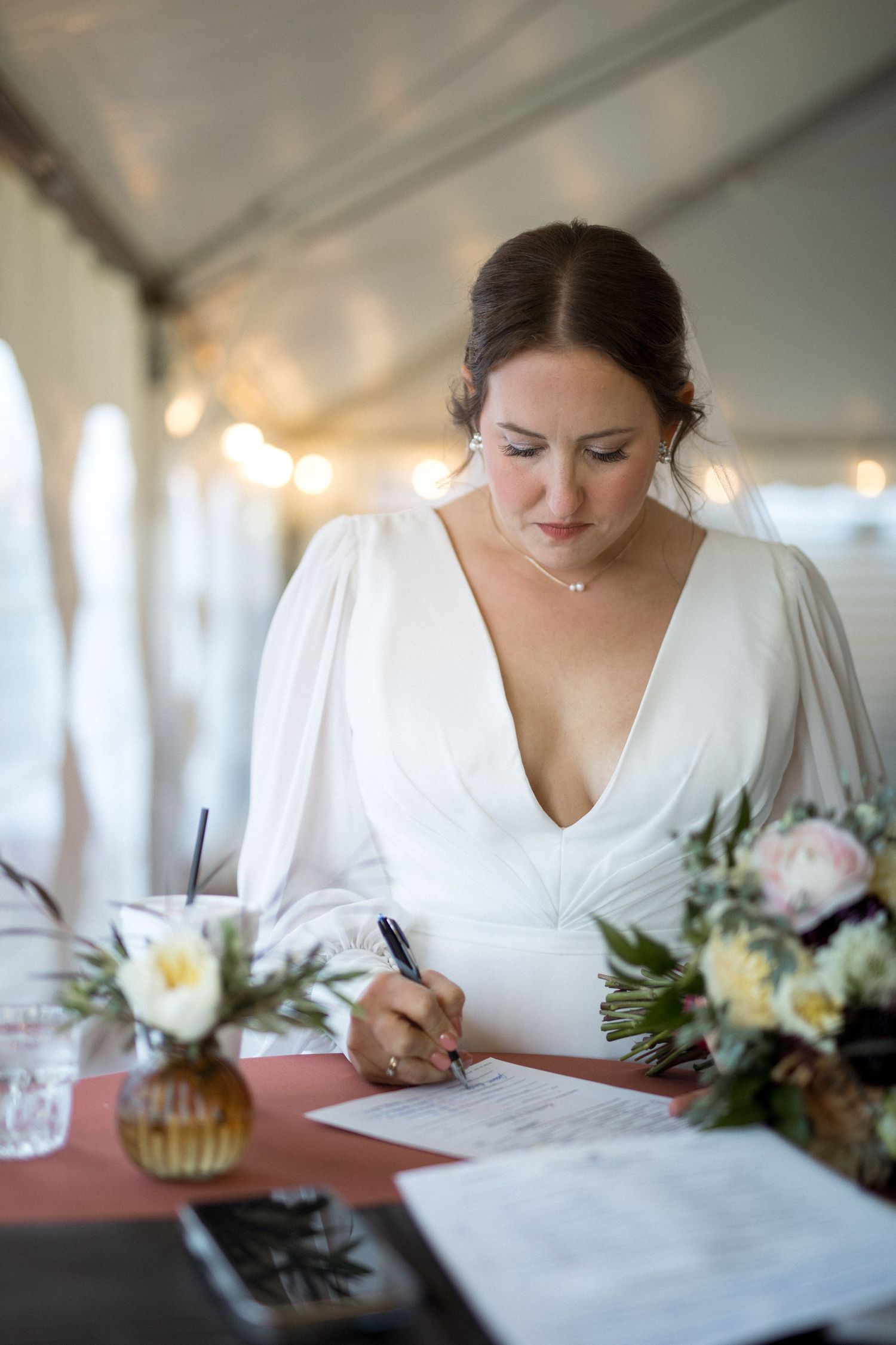 A bride signs wedding documents at a table decorated with floral arrangements under a white tent.