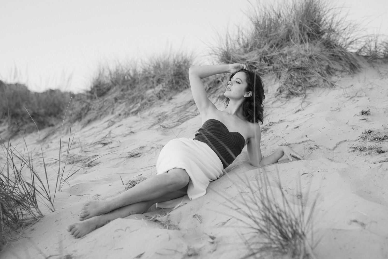 A series of black and white photos showing someone lounging and posing on a sandy beach with dune grass.