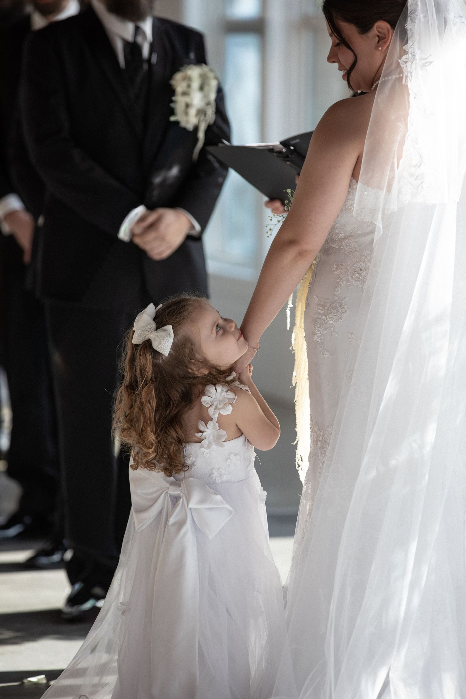 A flower girl in white dress holds the bride's hand during a wedding ceremony.
