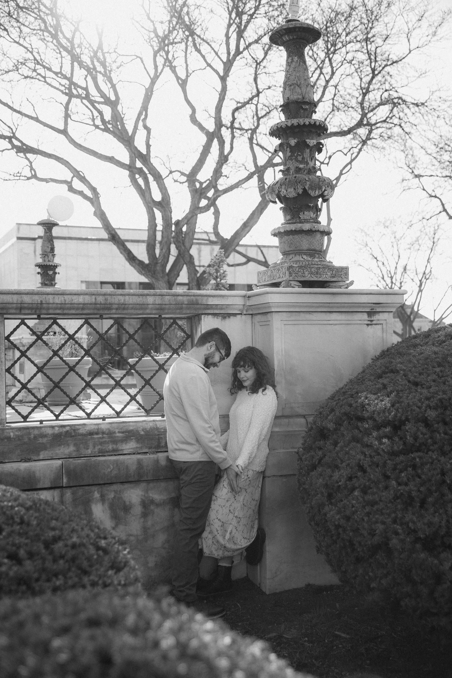 Black and white photo of a couple holding hands by an ornate stone fountain and lattice fence in a garden.
