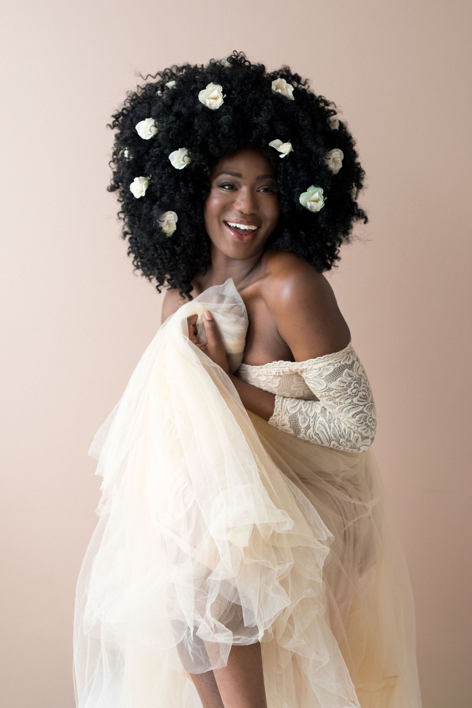 Smiling Black woman with white flowers in afro hair, wearing an elegant cream tulle gown, studio portrait.