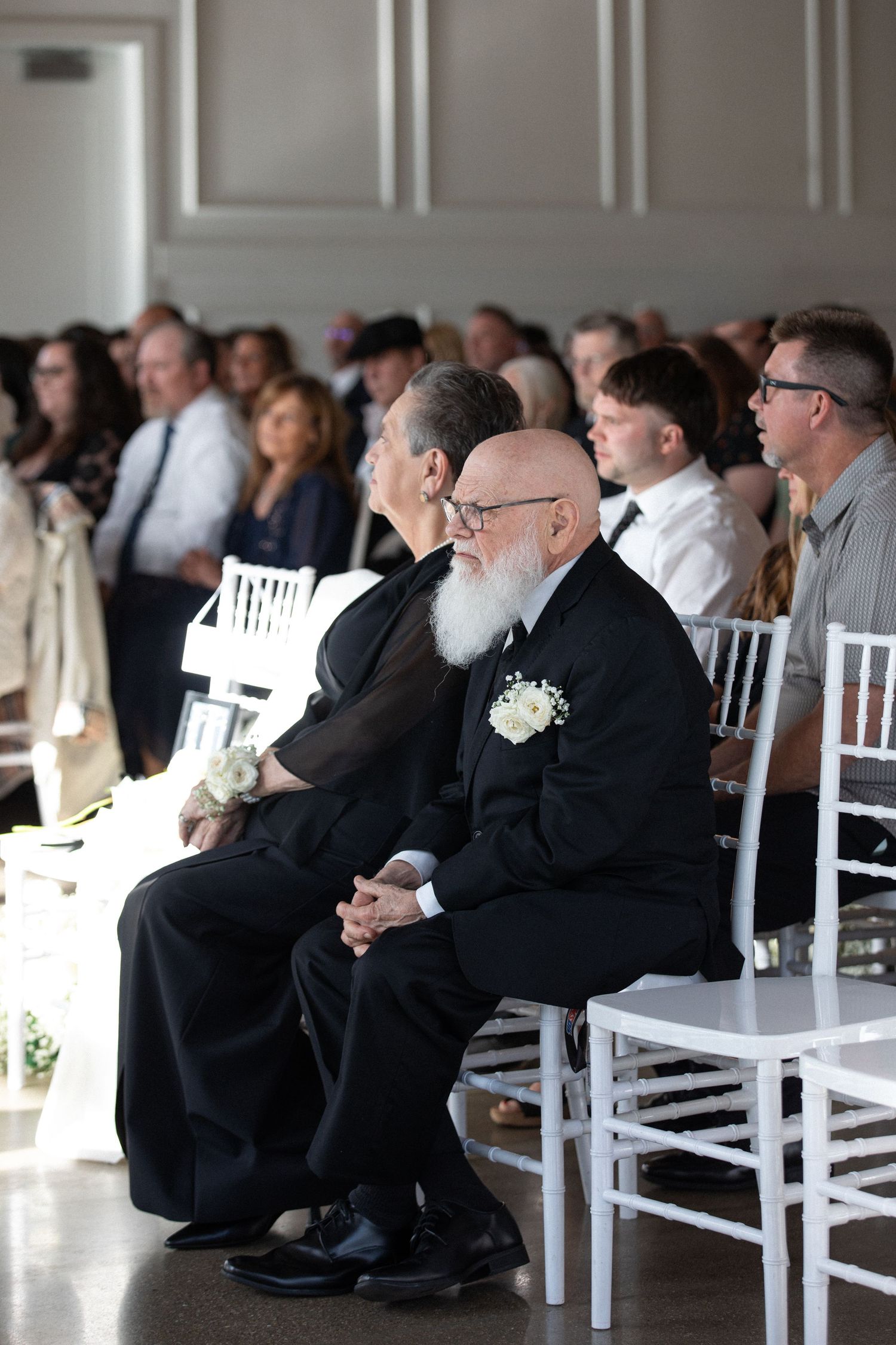 Wedding guests seated in white chairs during ceremony, including elderly man in black suit with white boutonniere.