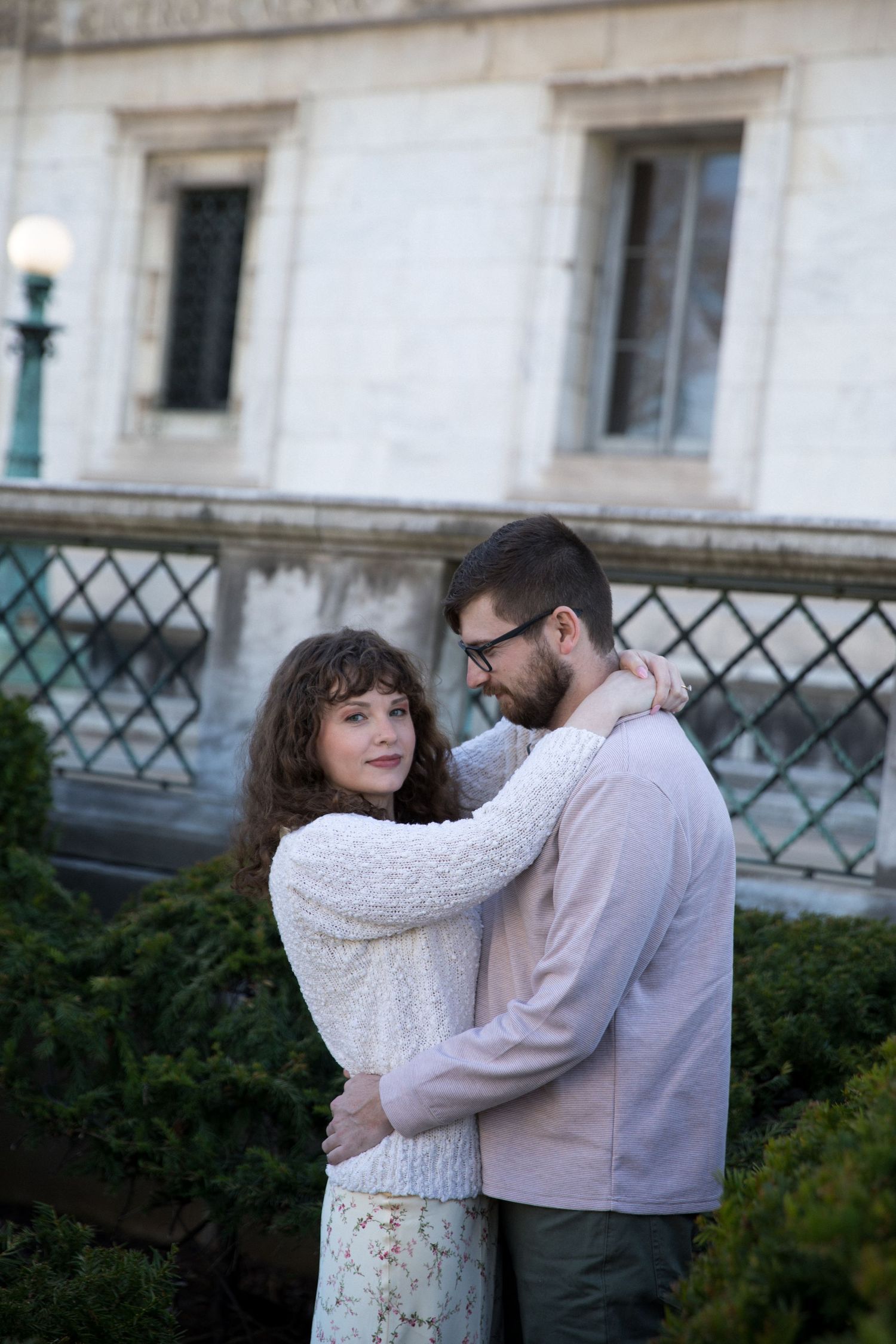 A couple embraces outdoors near a historic building, woman in white sweater smiling at camera, man in pink jacket wearing glasses.