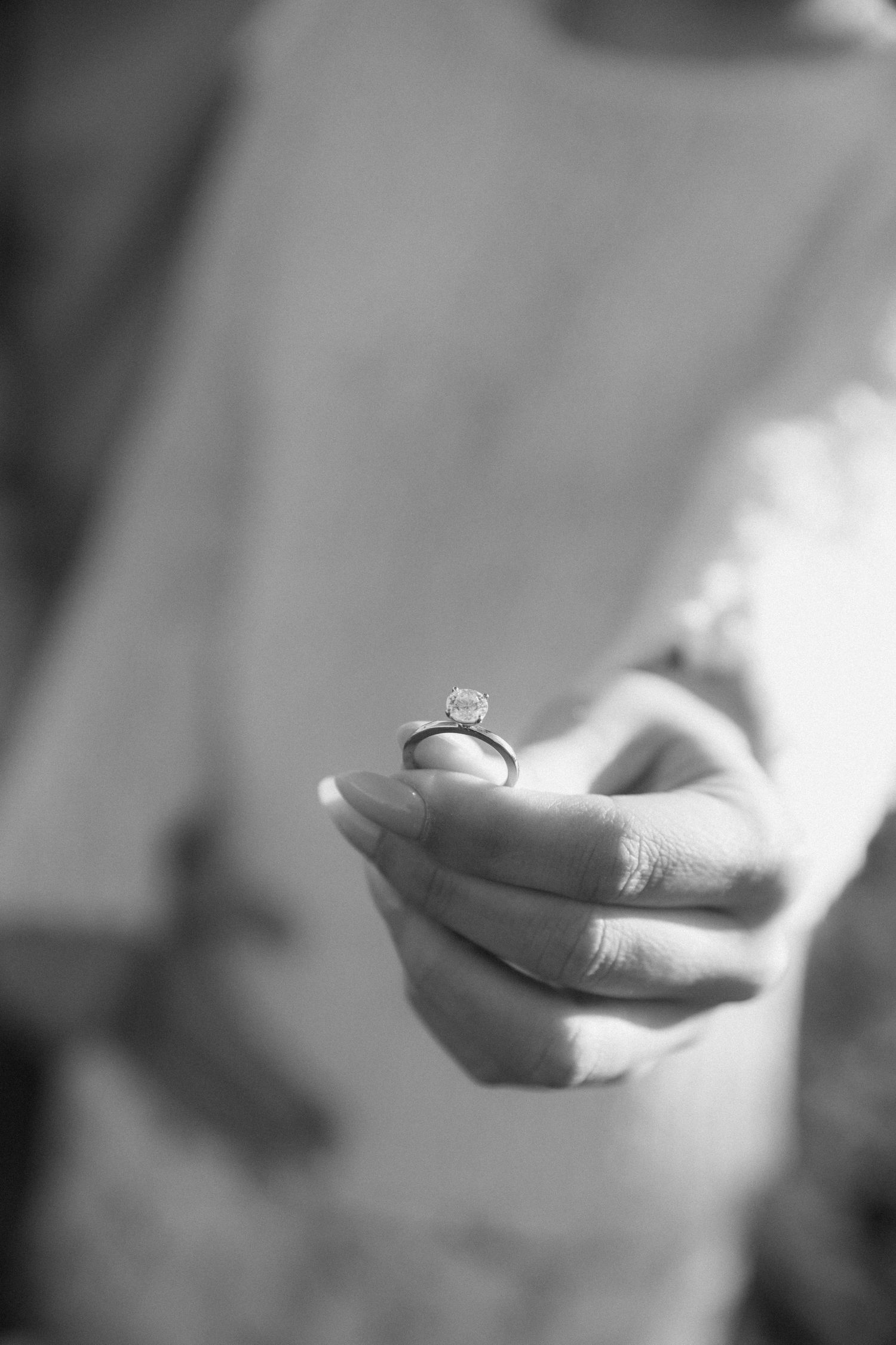 Black and white photo of a bride holding a diamond solitaire engagement ring between her fingers.