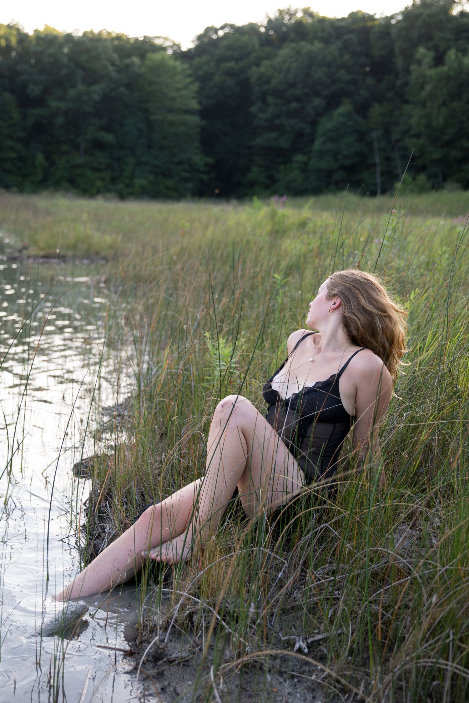 A person in a black dress lies gracefully among tall marsh grasses and shallow water at sunset.