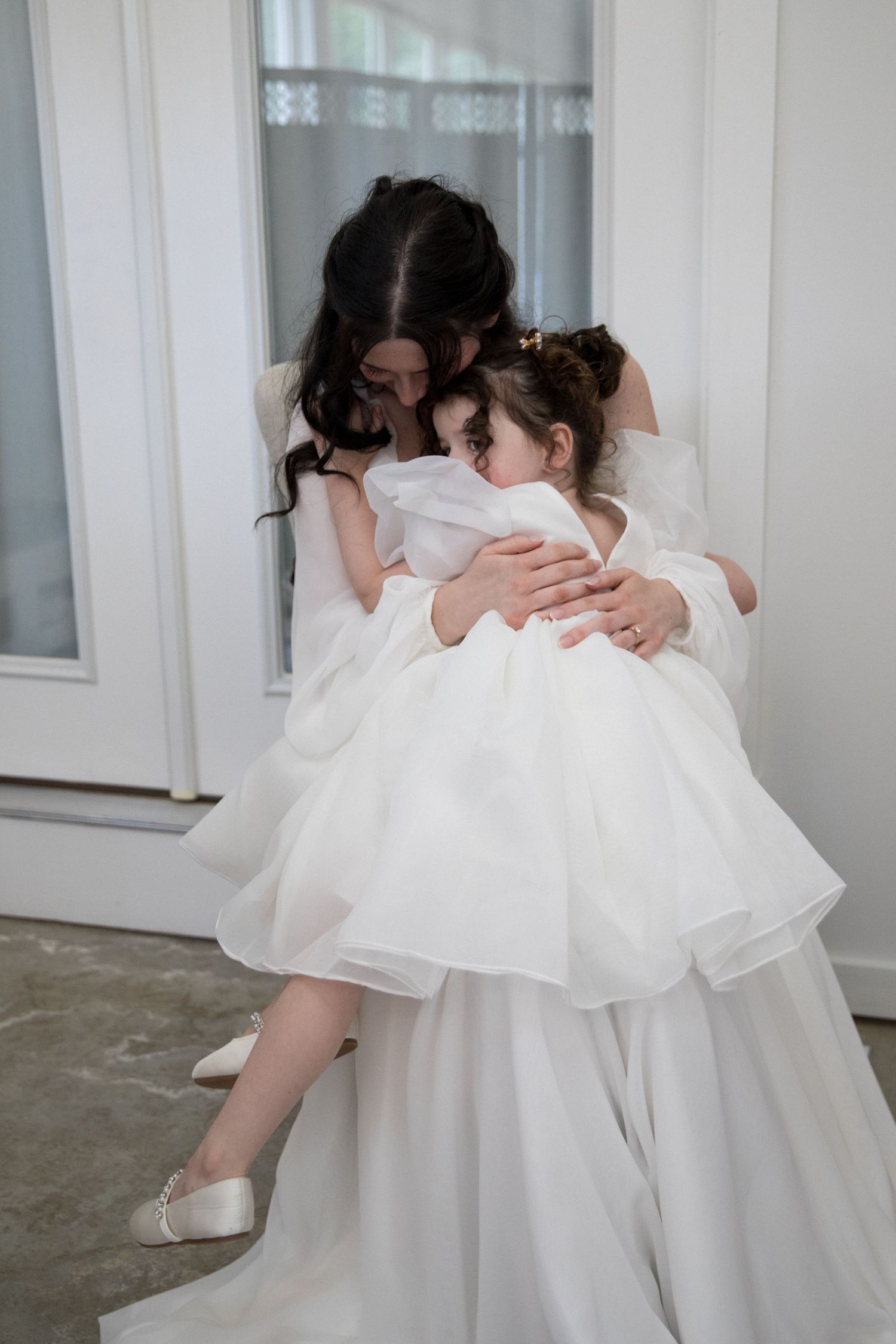 Bride in white wedding gown hugging a young flower girl in a matching white dress indoors.