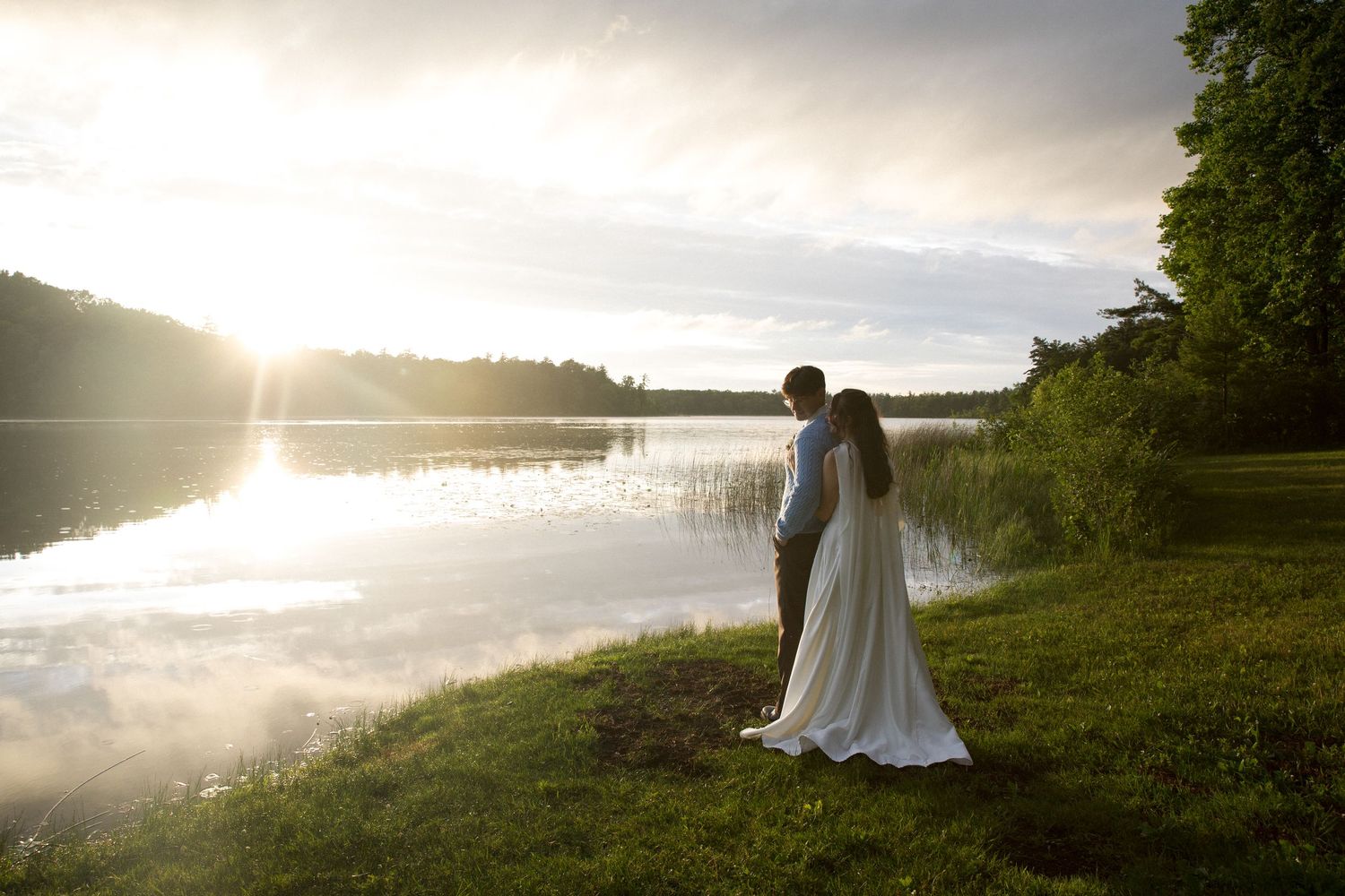 Bride and groom standing by a serene lake at sunset, surrounded by lush green grass and trees.