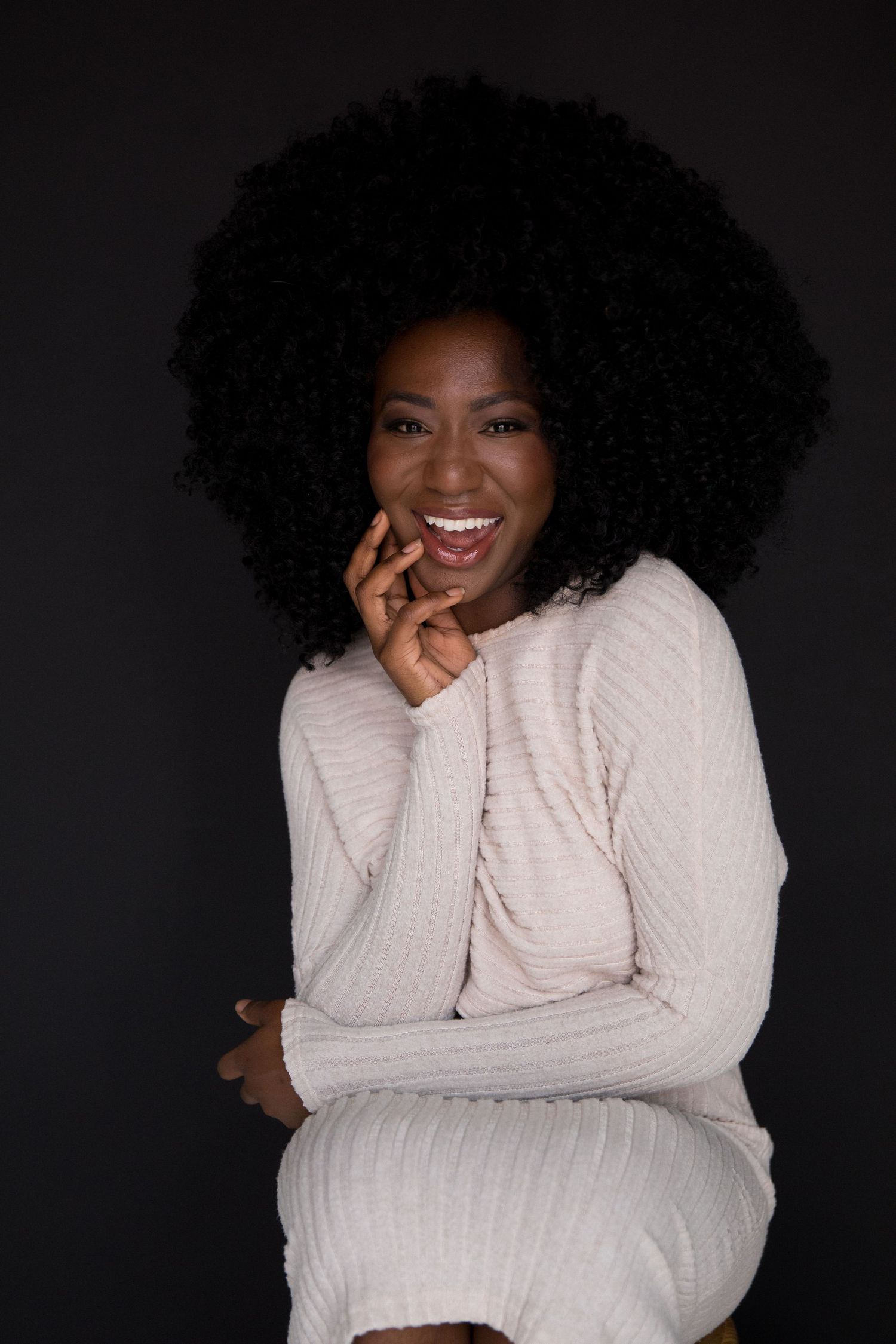 Smiling Black woman with natural afro hair wearing a light pink ribbed sweater, posing against a dark background.