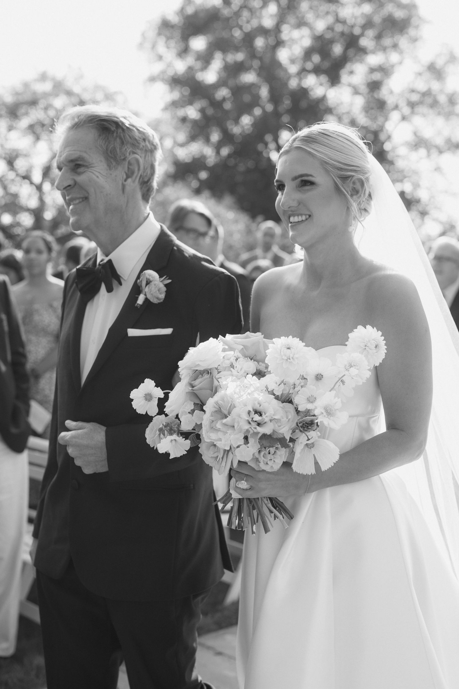 A couple in wedding attire poses outdoors with white floral bouquet.