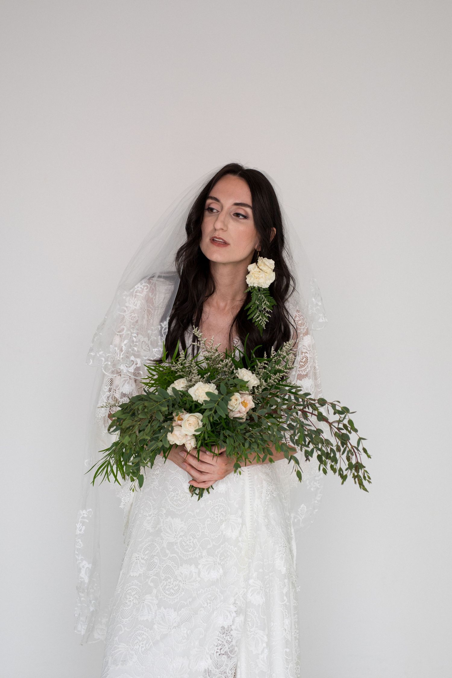 Bride in white lace gown and veil holds greenery bouquet with cream roses, posing against white background.