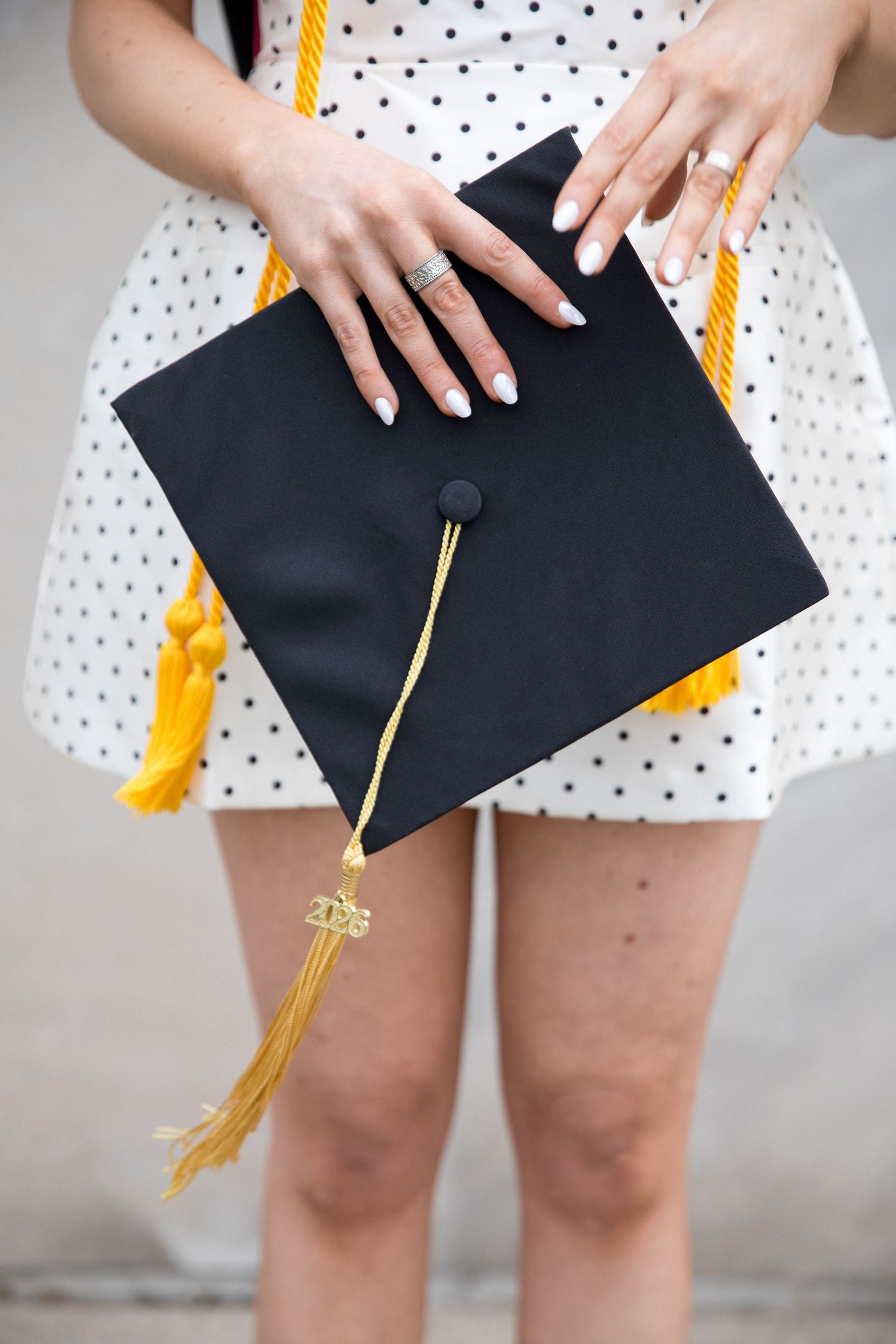 Woman in polka dot dress holding black graduation cap with gold tassel, showcasing white manicured nails and ring.