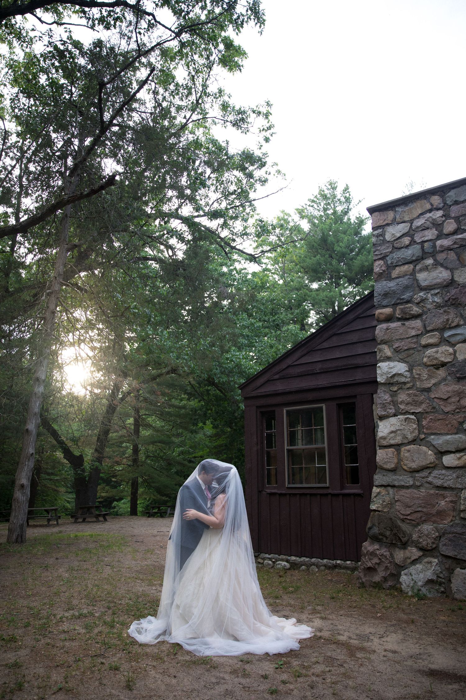 Bride and groom share a kiss under a flowing veil beside a rustic stone building in a wooded setting.