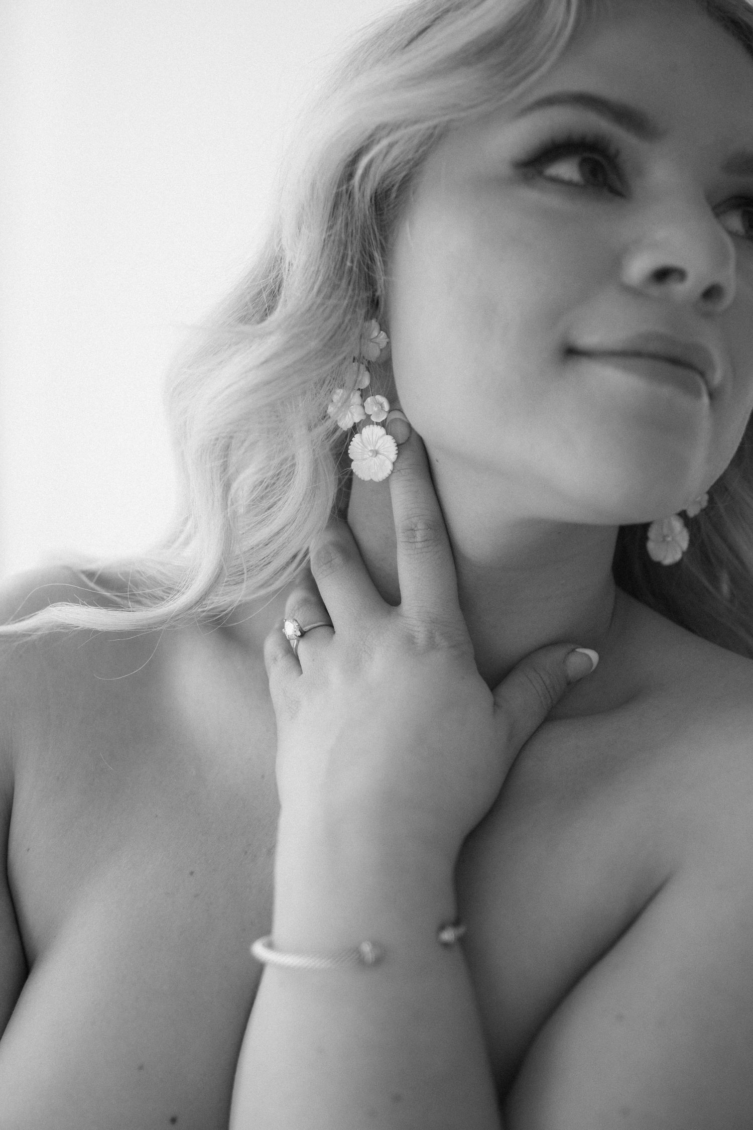 Black and white photo of a smiling blonde woman touching floral earring, wearing a bracelet and ring.