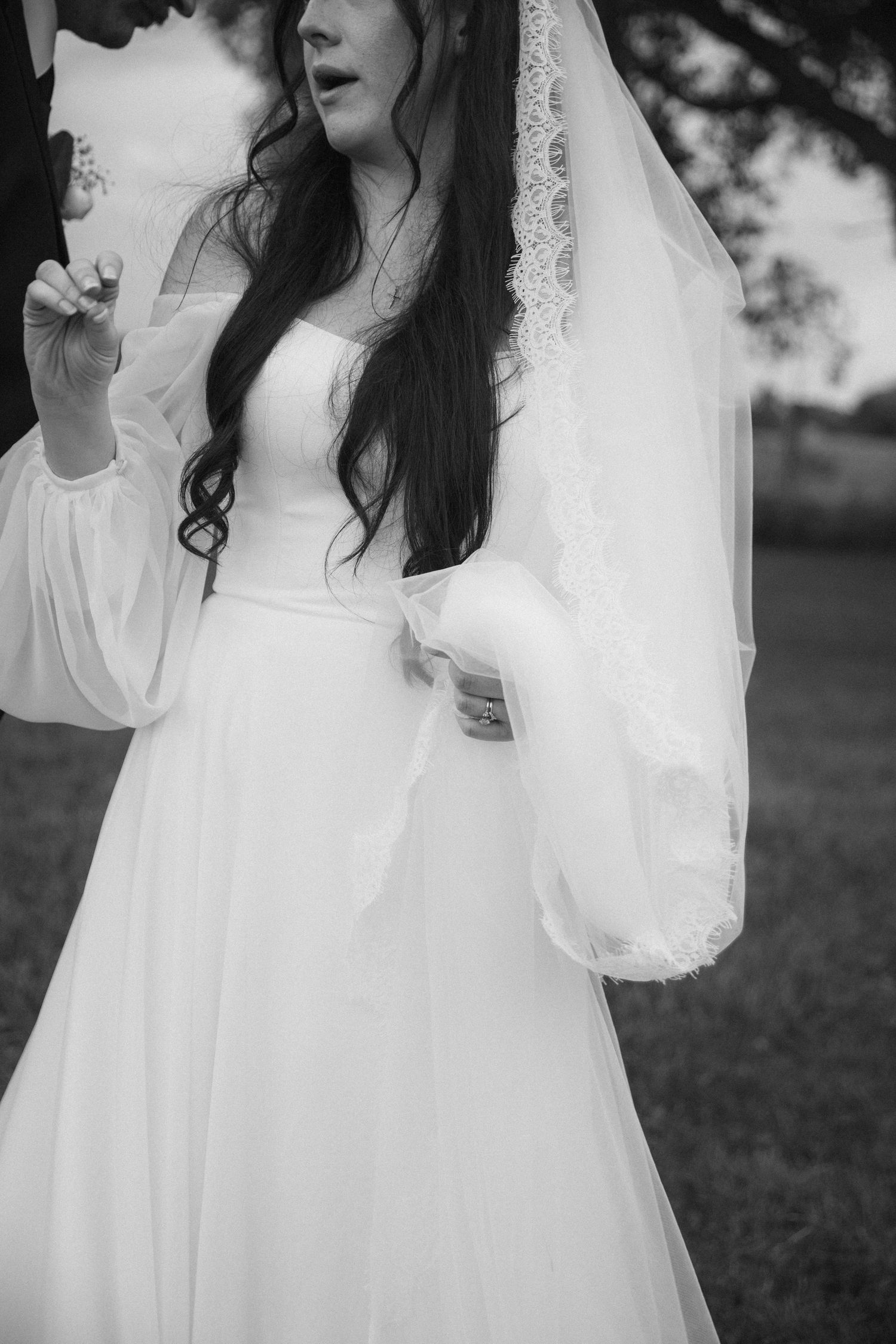 Black and white photo of bride in elegant white dress with lace veil and puff sleeves outdoors on wedding day.
