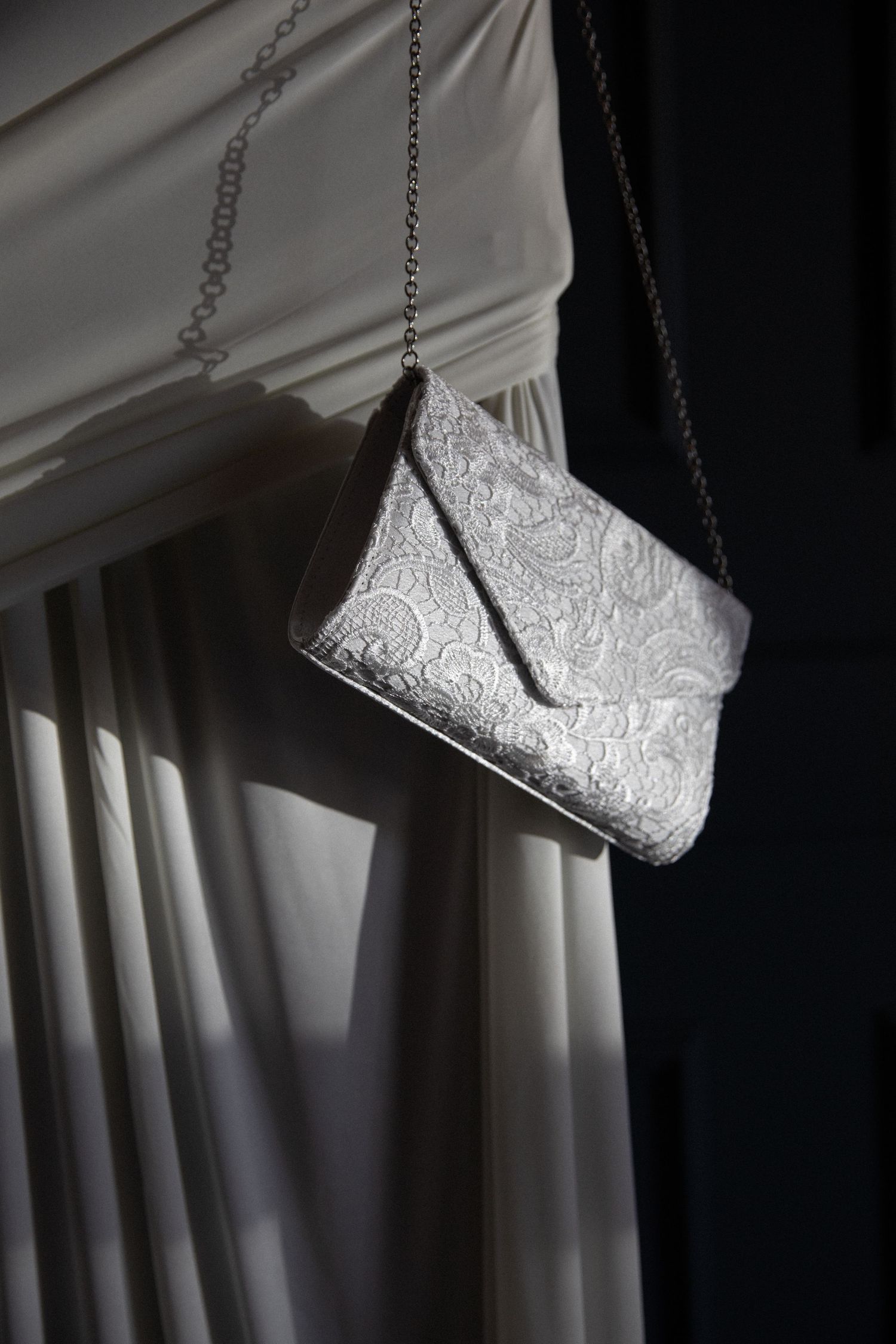Silver lace embroidered clutch purse hanging on chain strap against white draped curtain.