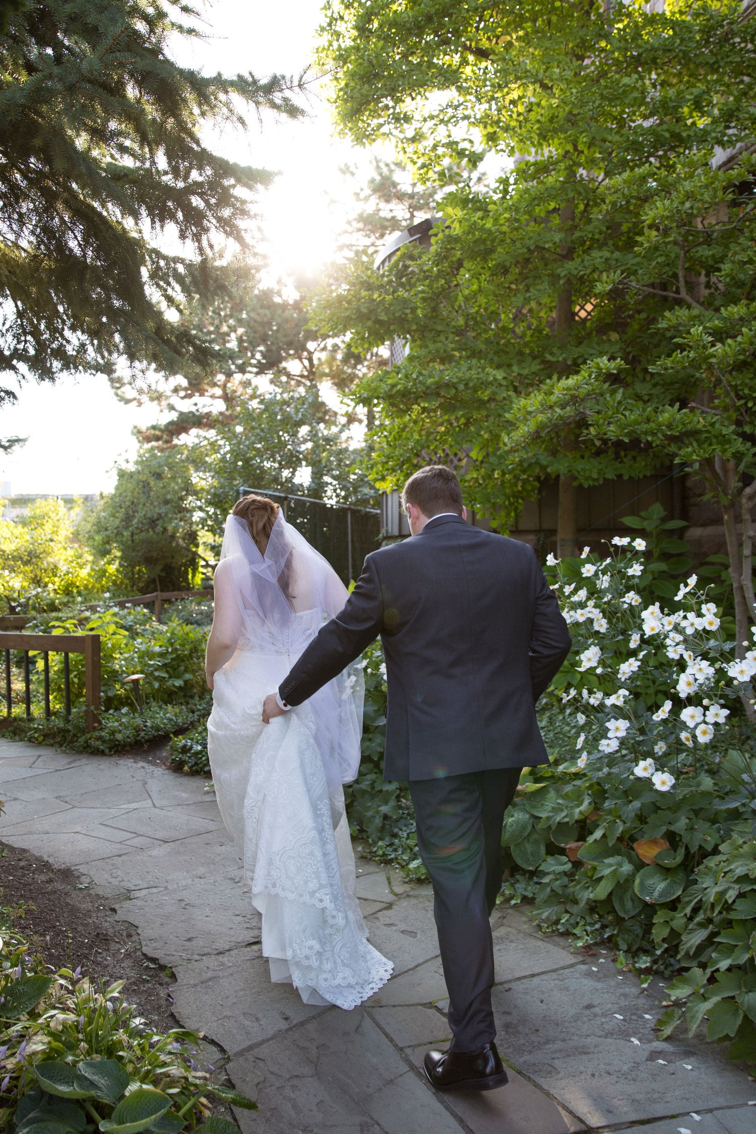 A couple walks along a garden path surrounded by lush greenery and white flowers on a sunny afternoon.