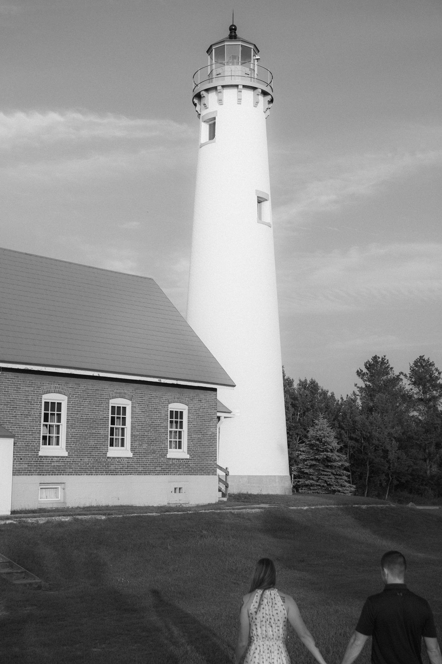 A tall white lighthouse stands next to a brick building as two people view it from the lawn in black and white.