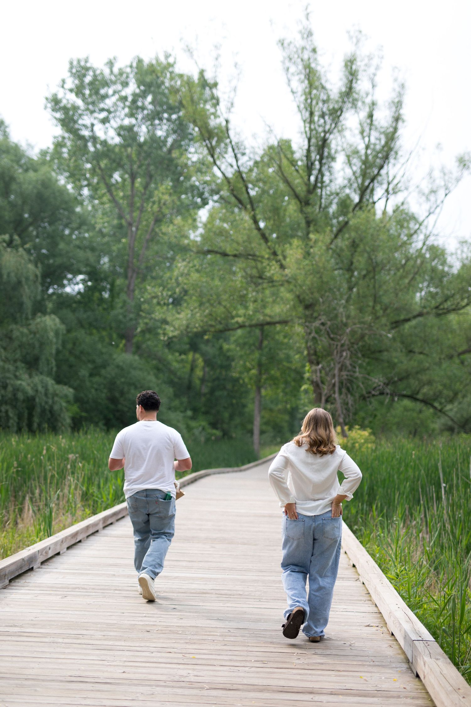 Two people walking away on a wooden boardwalk path surrounded by lush green trees and tall grass.
