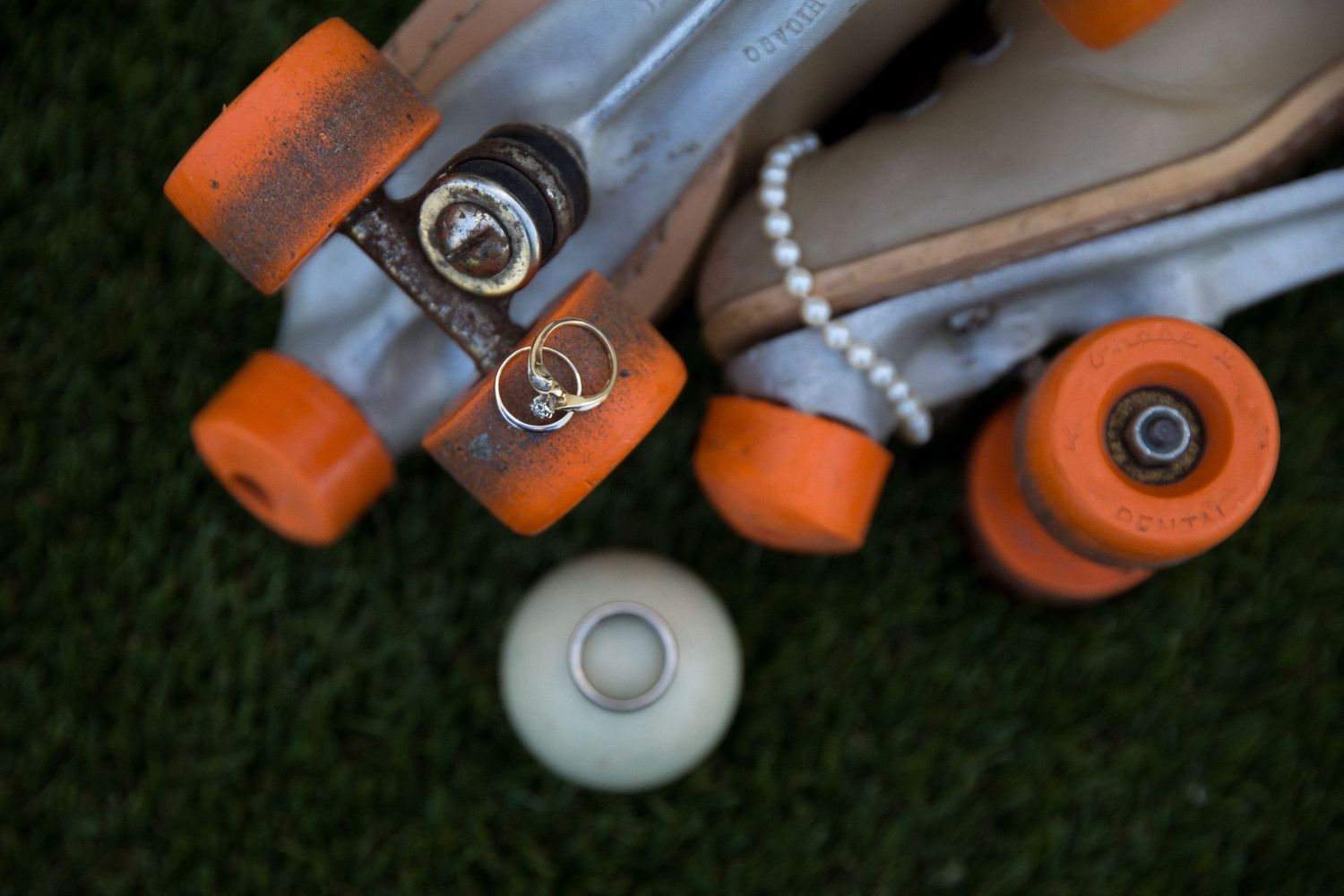 Wedding rings resting on orange roller skate wheels on green grass with pearl bracelet detail.