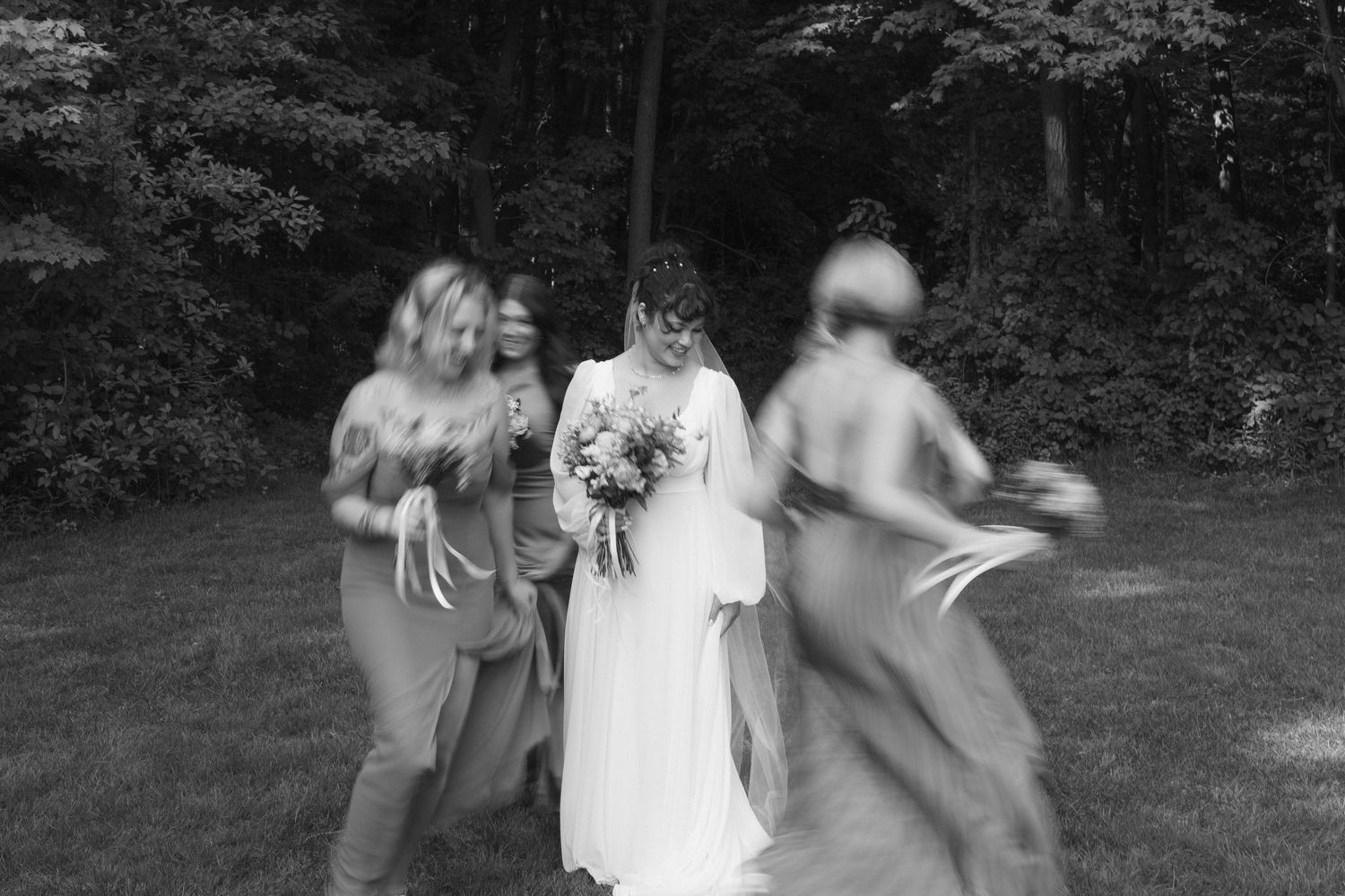 Black and white photo of a bride holding a bouquet surrounded by bridesmaids in flowing dresses outdoors.