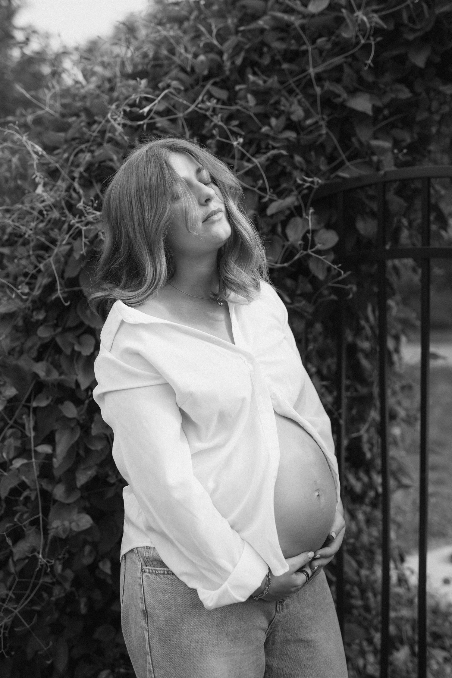 Black and white photo of a pregnant woman in an open white shirt and jeans, cradling her baby bump outdoors.