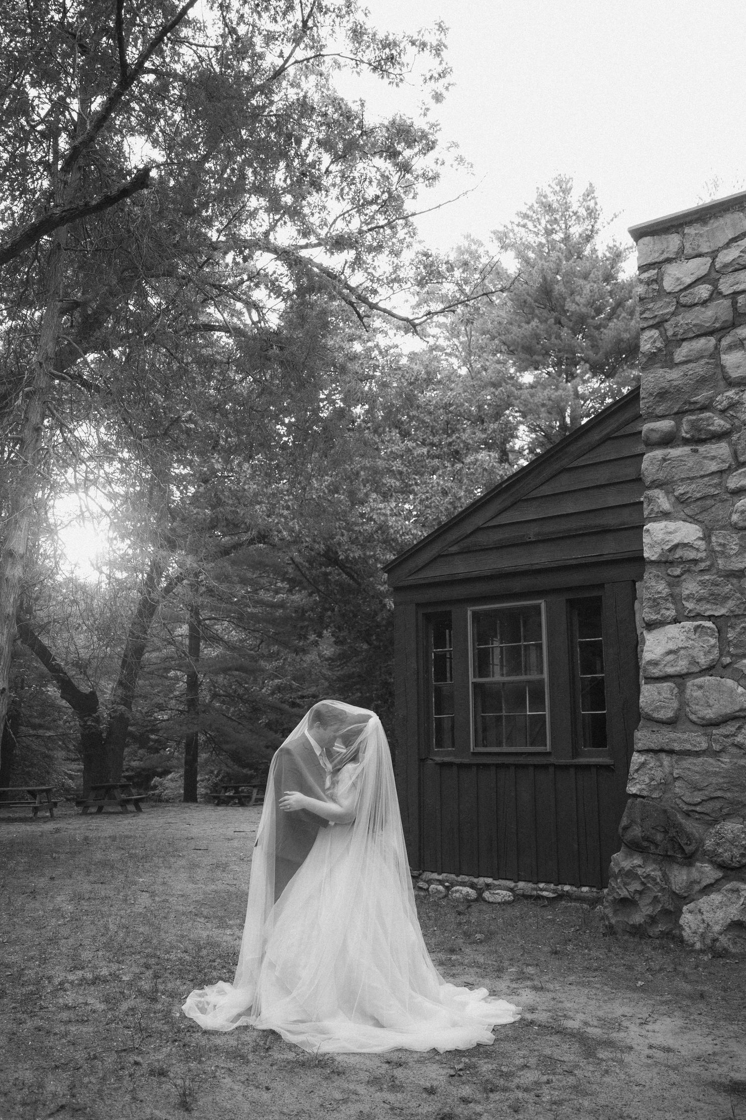 Black and white photo of a bride covered in a flowing veil outdoors near a rustic stone and wood cabin surrounded by trees.