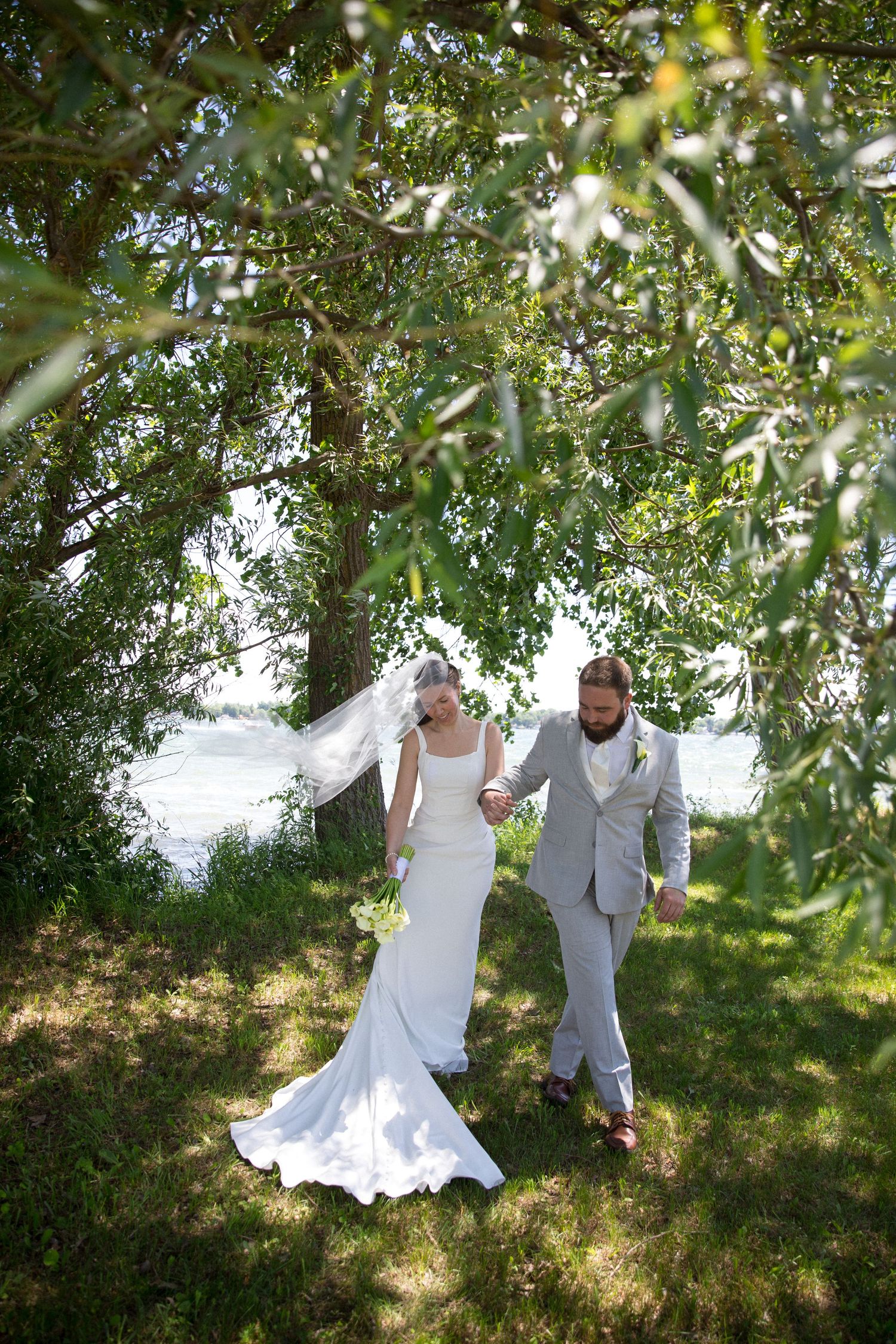 Bride and groom walk hand-in-hand under a willow tree by a lake, bride wearing a white gown with veil.