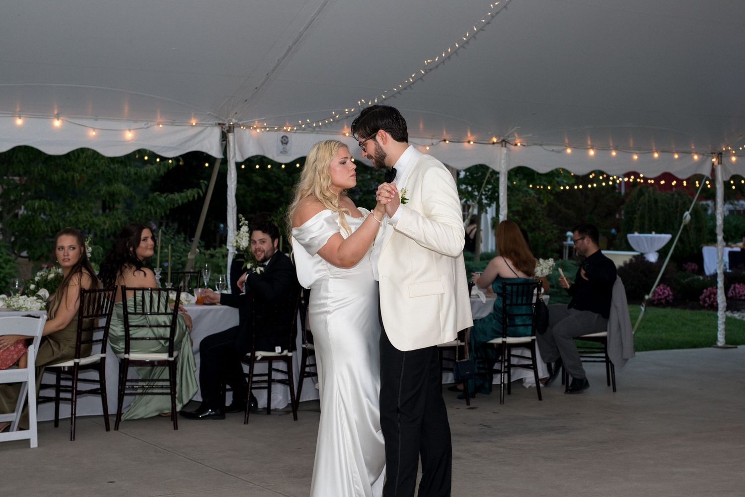 Couples dancing under string lights at an outdoor wedding reception with a white tent canopy.