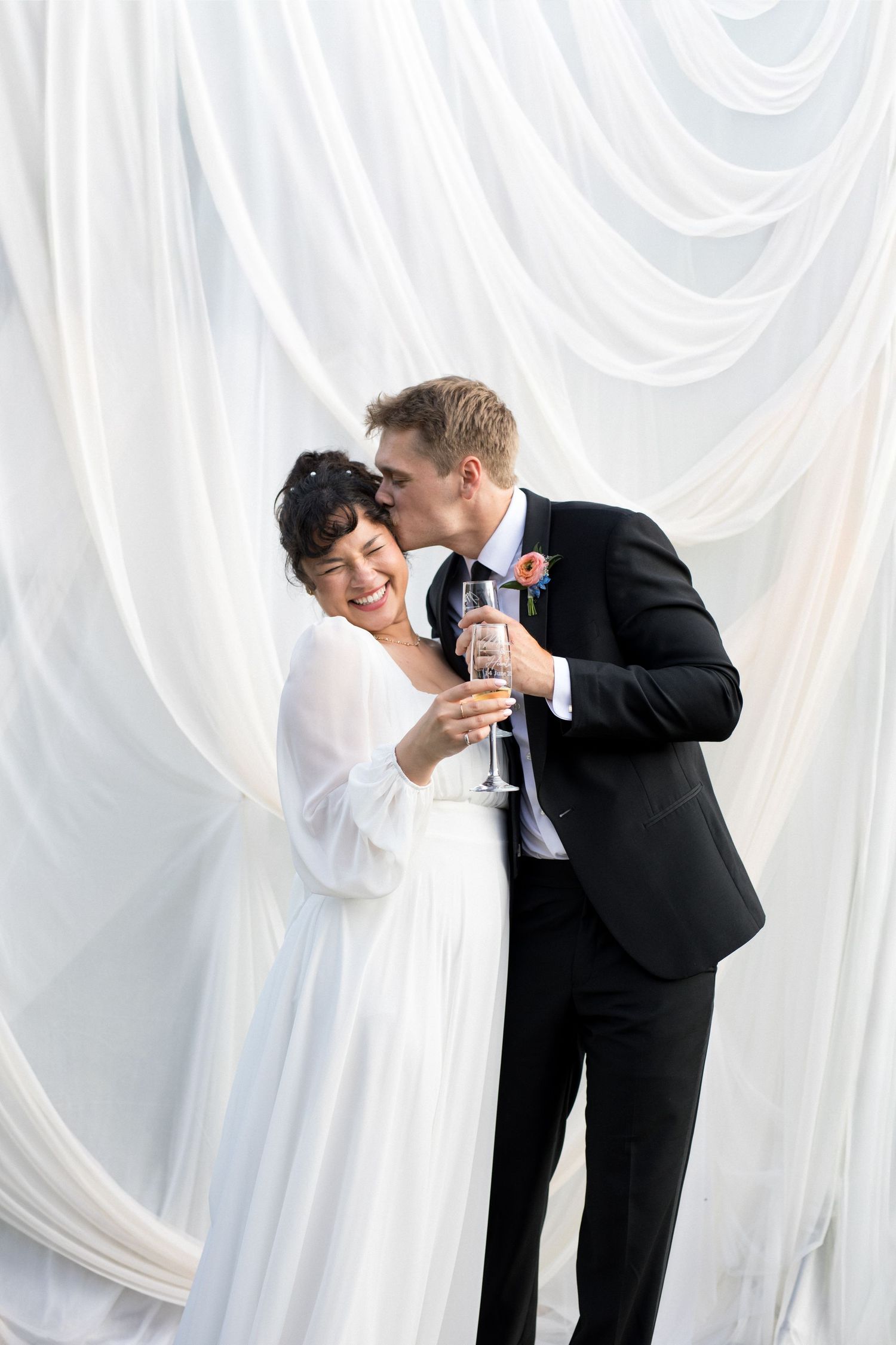 A bride and groom in formal attire embrace and hold a microphone together, standing in front of a white draped backdrop.