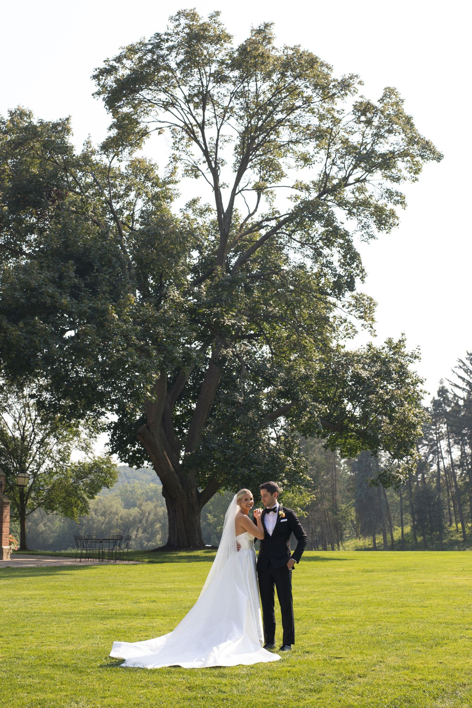 Wedding couple stands together on a sunlit grassy hill beneath a majestic oak tree during an outdoor ceremony.